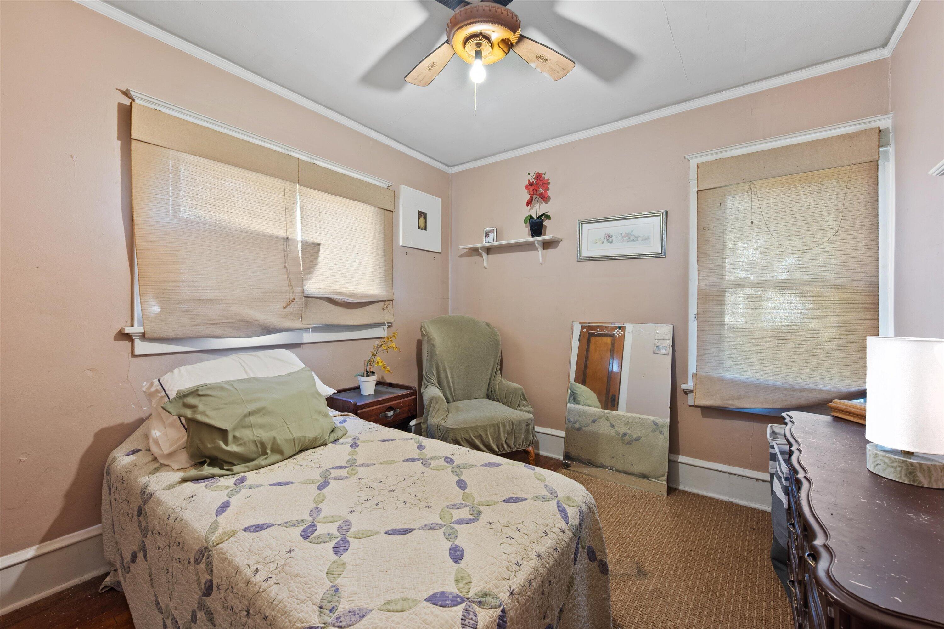 Bedroom with a patterned quilt-covered bed, green chair, beige blinds, and ceiling fan.
