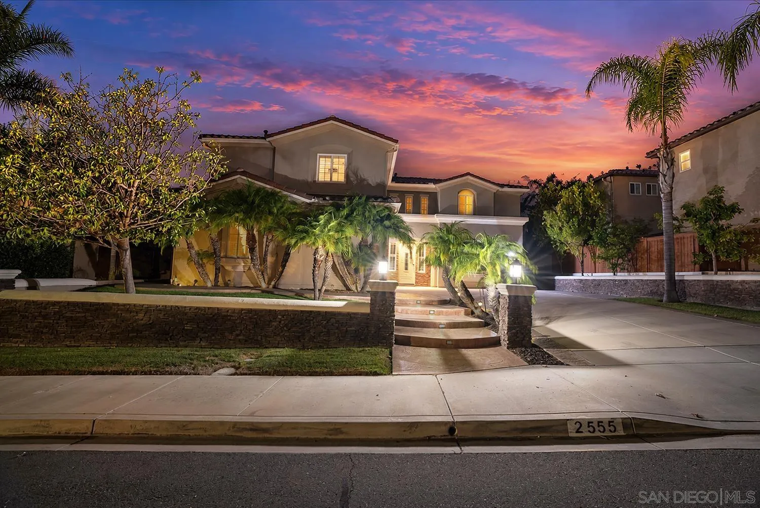 Two-story stucco home at dusk, framed by palm trees and a low stone wall.