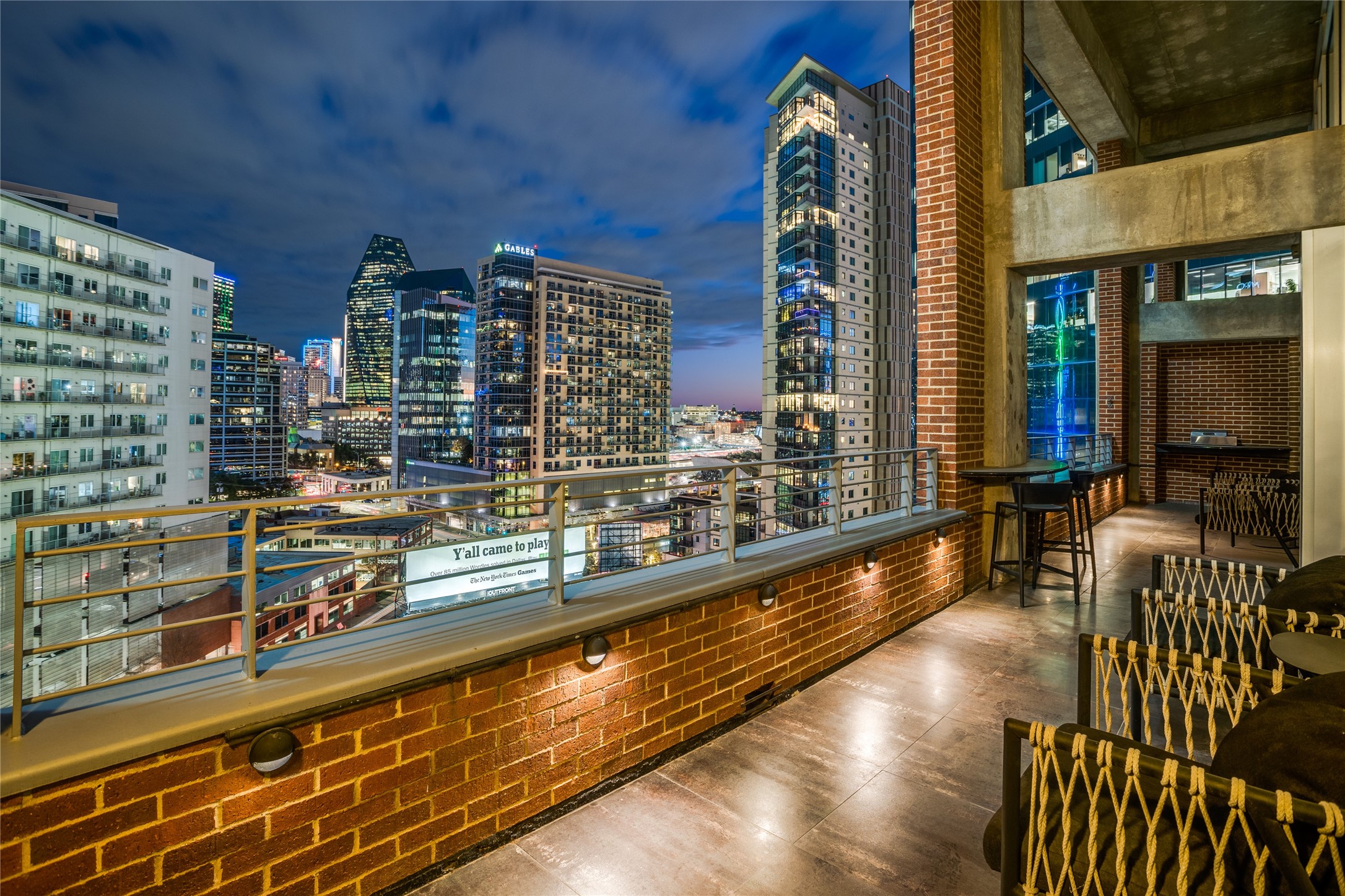 Furnished brick balcony overlooking the illuminated Dallas skyline, with outdoor seating and a built-in fireplace.
