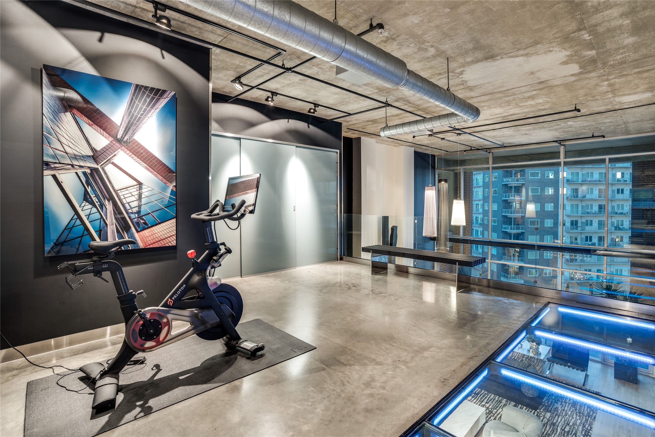 Condominium fitness room with Peloton bikes, exposed ductwork, and a blue-lit glass floor section.