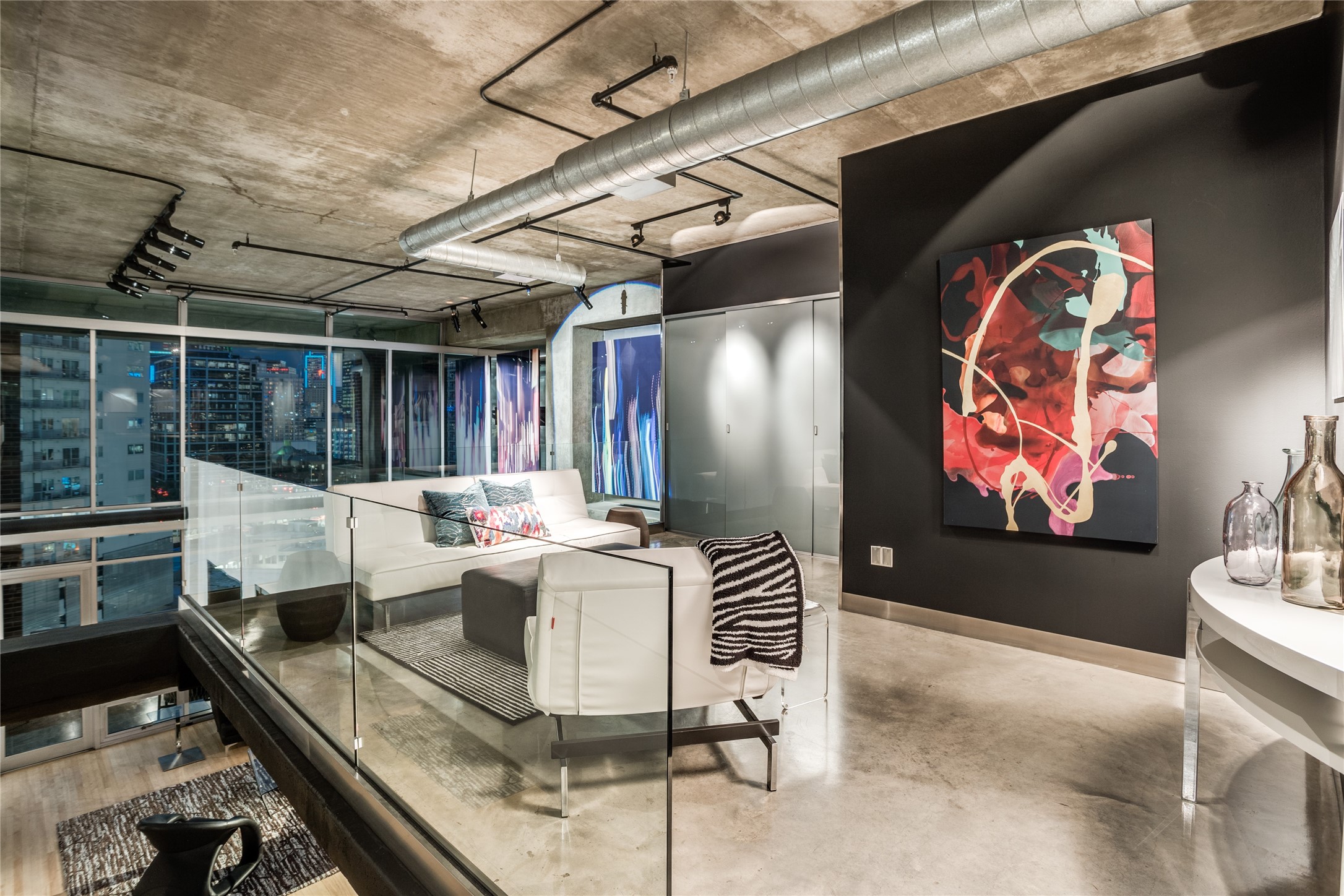 Modern loft living room with white seating, exposed concrete ceiling, and nighttime Dallas skyline views.