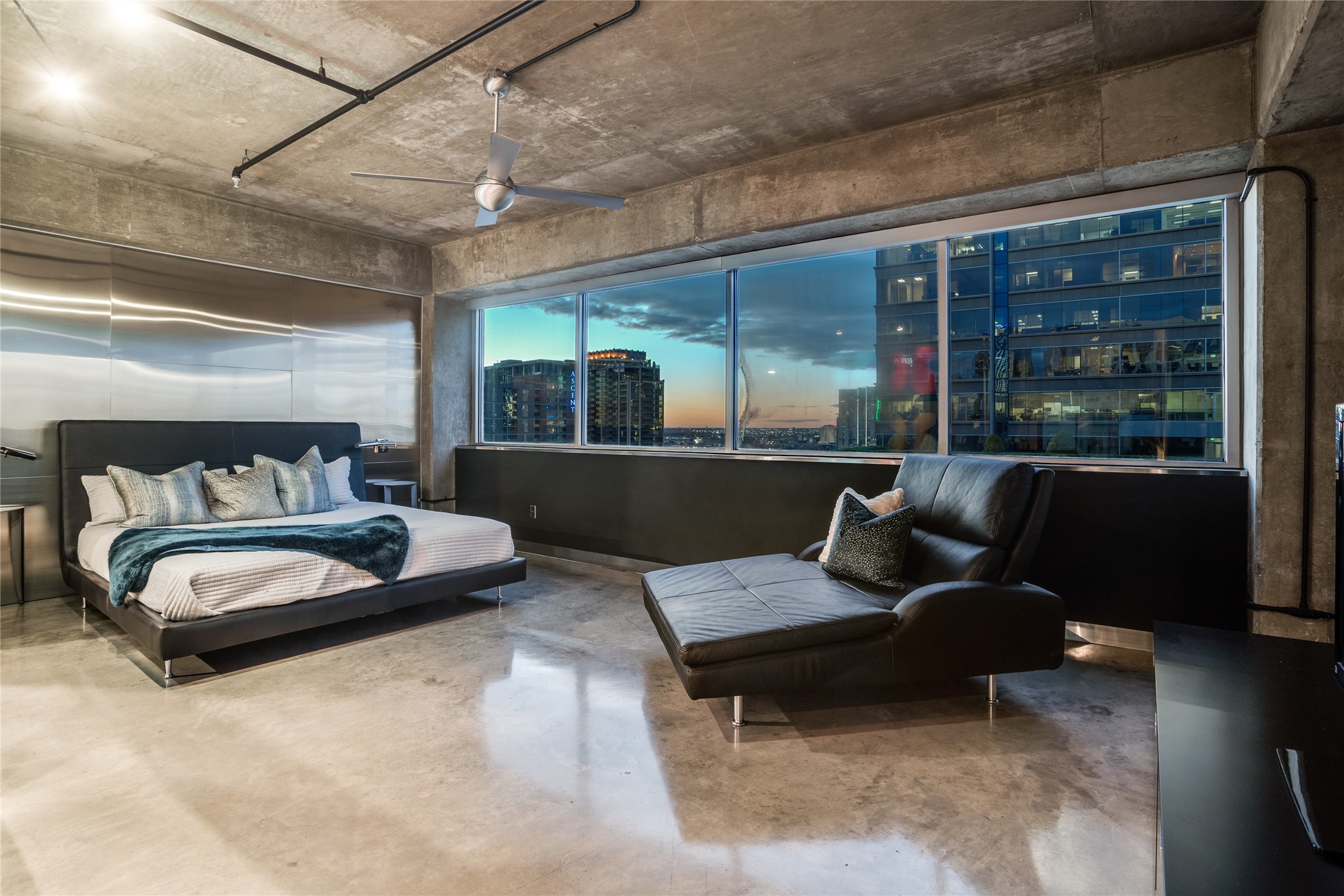 Modern bedroom with a platform bed, leather chaise, exposed concrete, and downtown views at dusk.