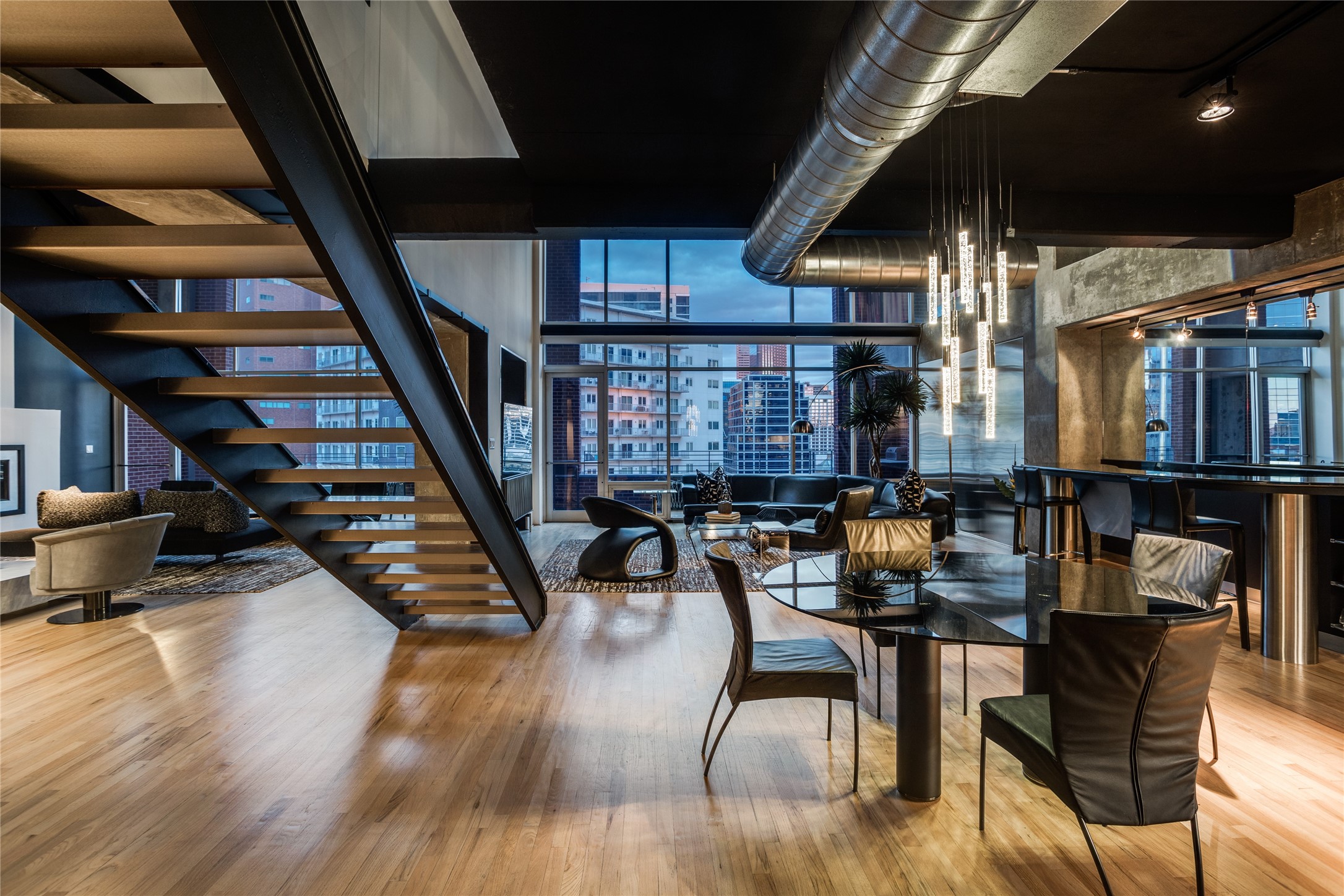 Loft living and dining area with an open staircase, hardwood floors, and city views through large windows.