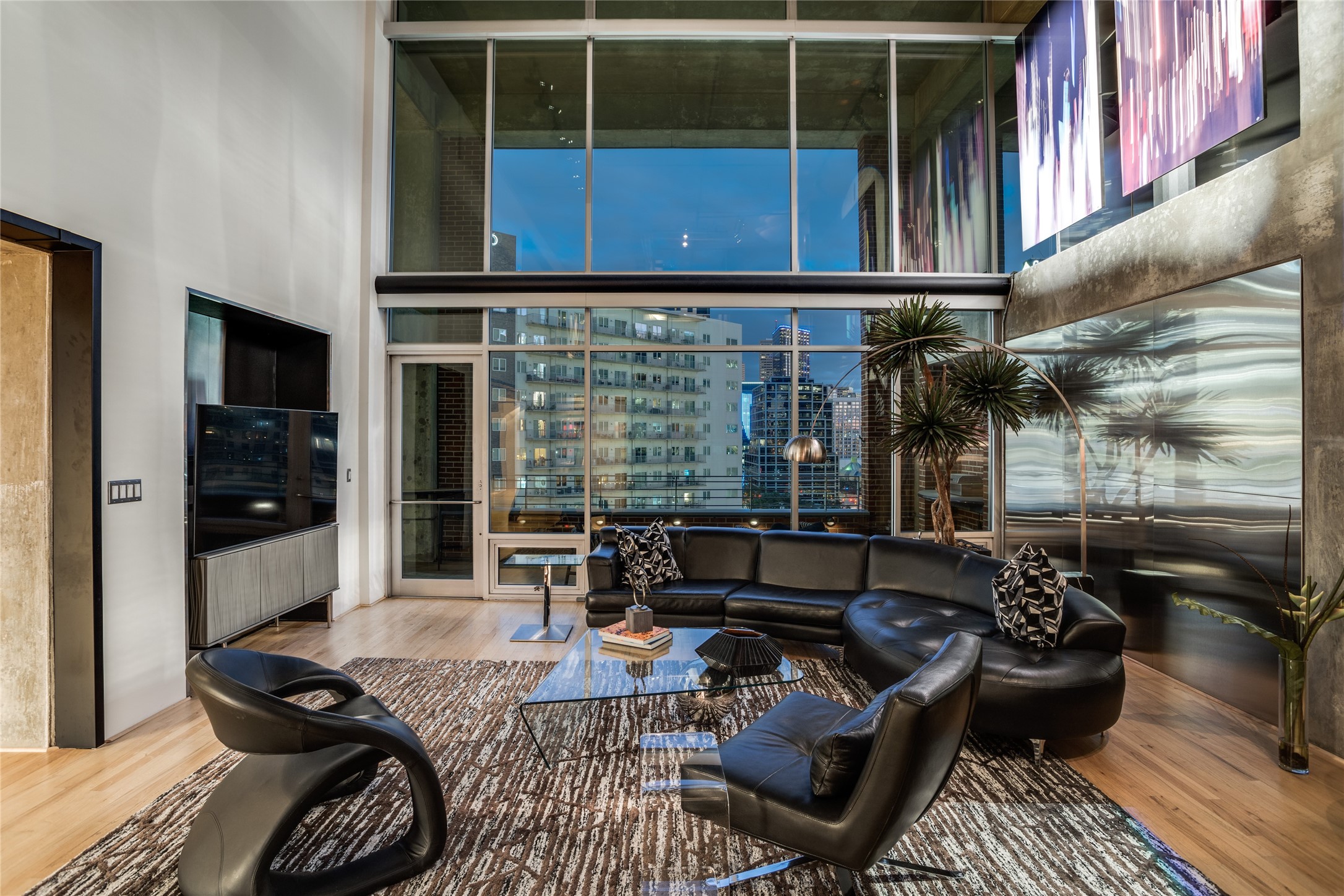 Modern condo living room with black leather seating and floor-to-ceiling windows overlooking downtown at dusk.