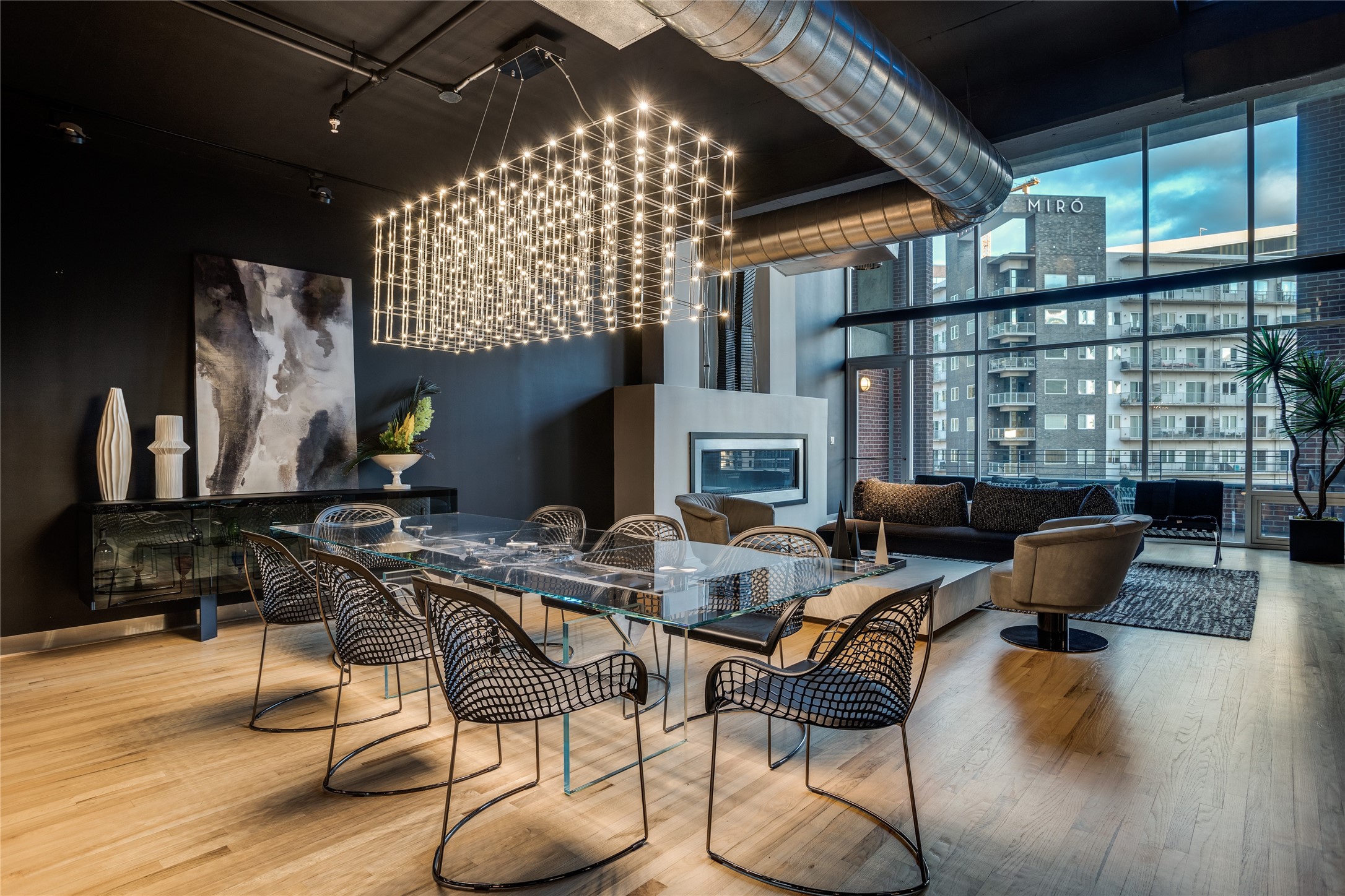 Open-plan condo living and dining area with a glass table, sculptural chandelier, fireplace, and floor-to-ceiling windows.