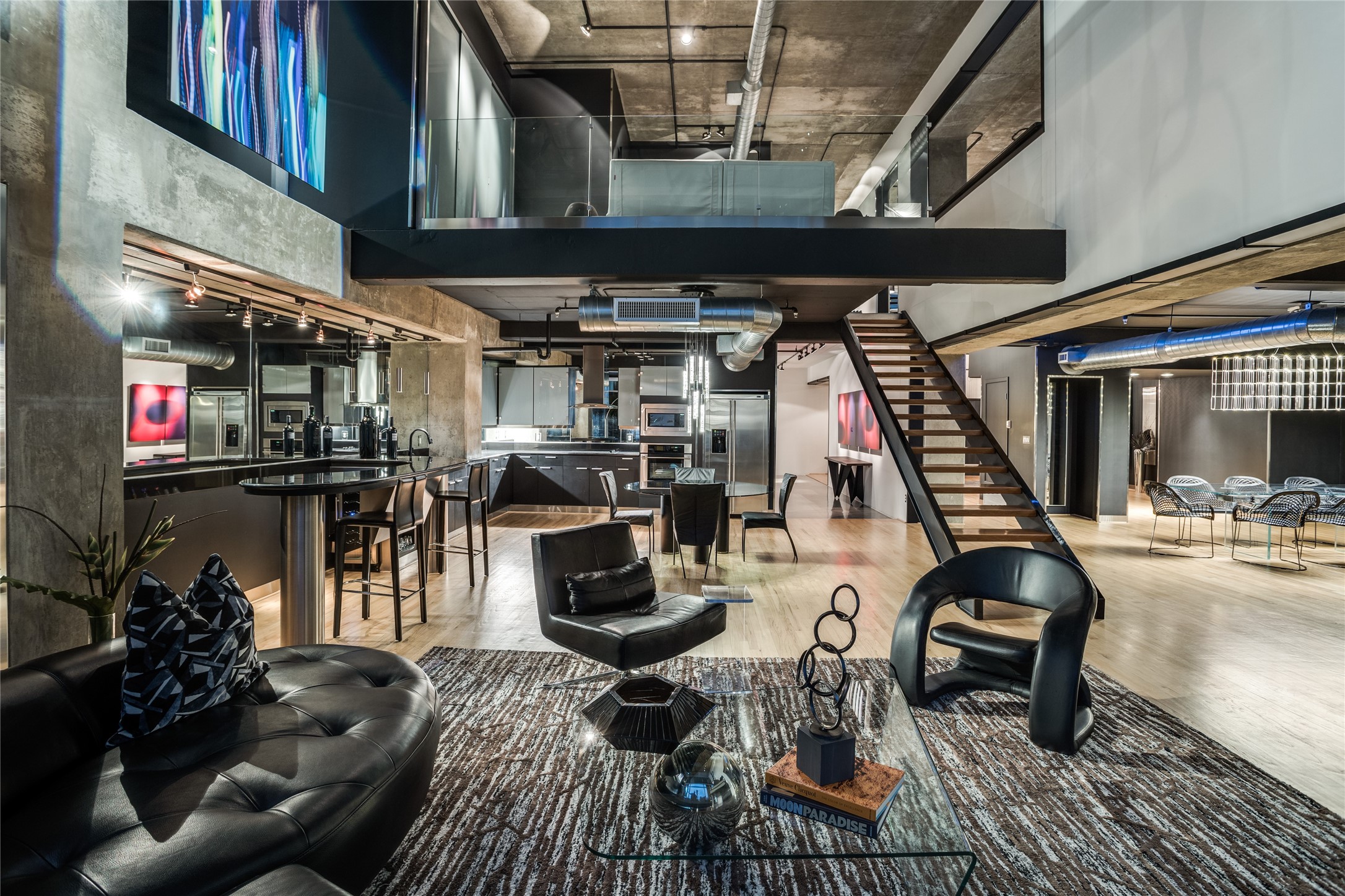 Loft living room with black leather seating, an open kitchen, exposed concrete ceiling, and mezzanine staircase.