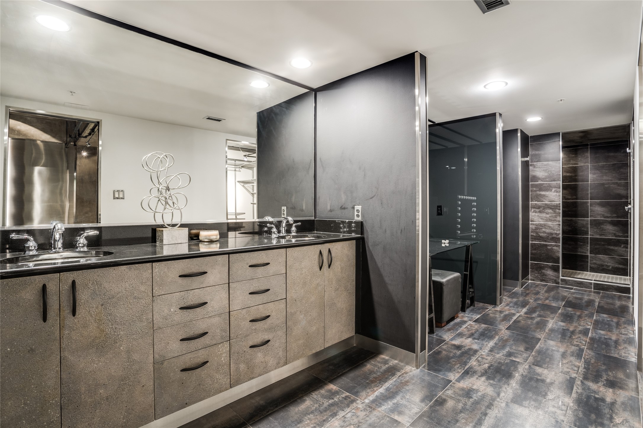 Modern bathroom with a long double vanity and glass shower lined in dark tile.