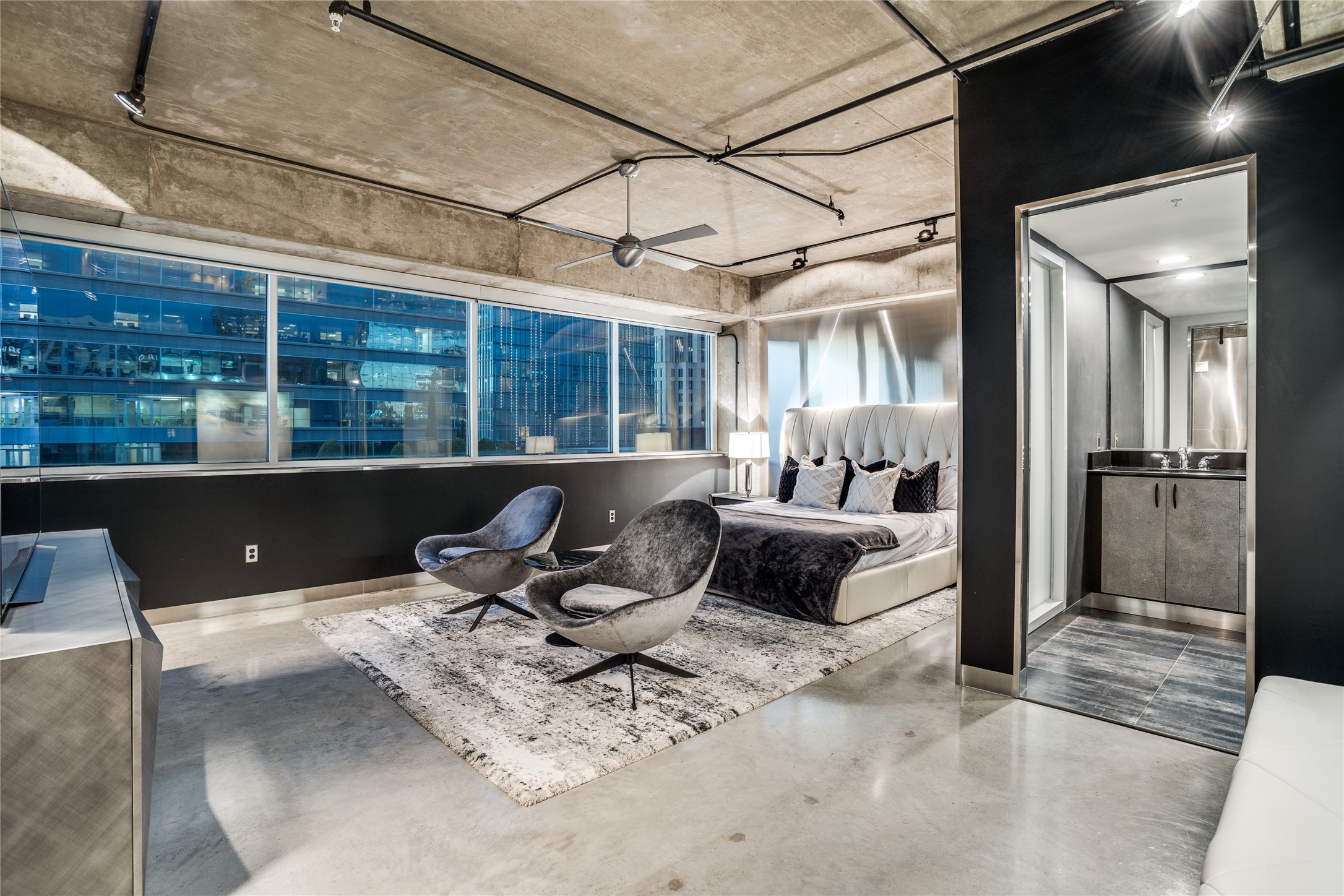 Modern condo bedroom with sculptural lounge chairs, floor-to-ceiling city-view windows, and an open bathroom vanity.