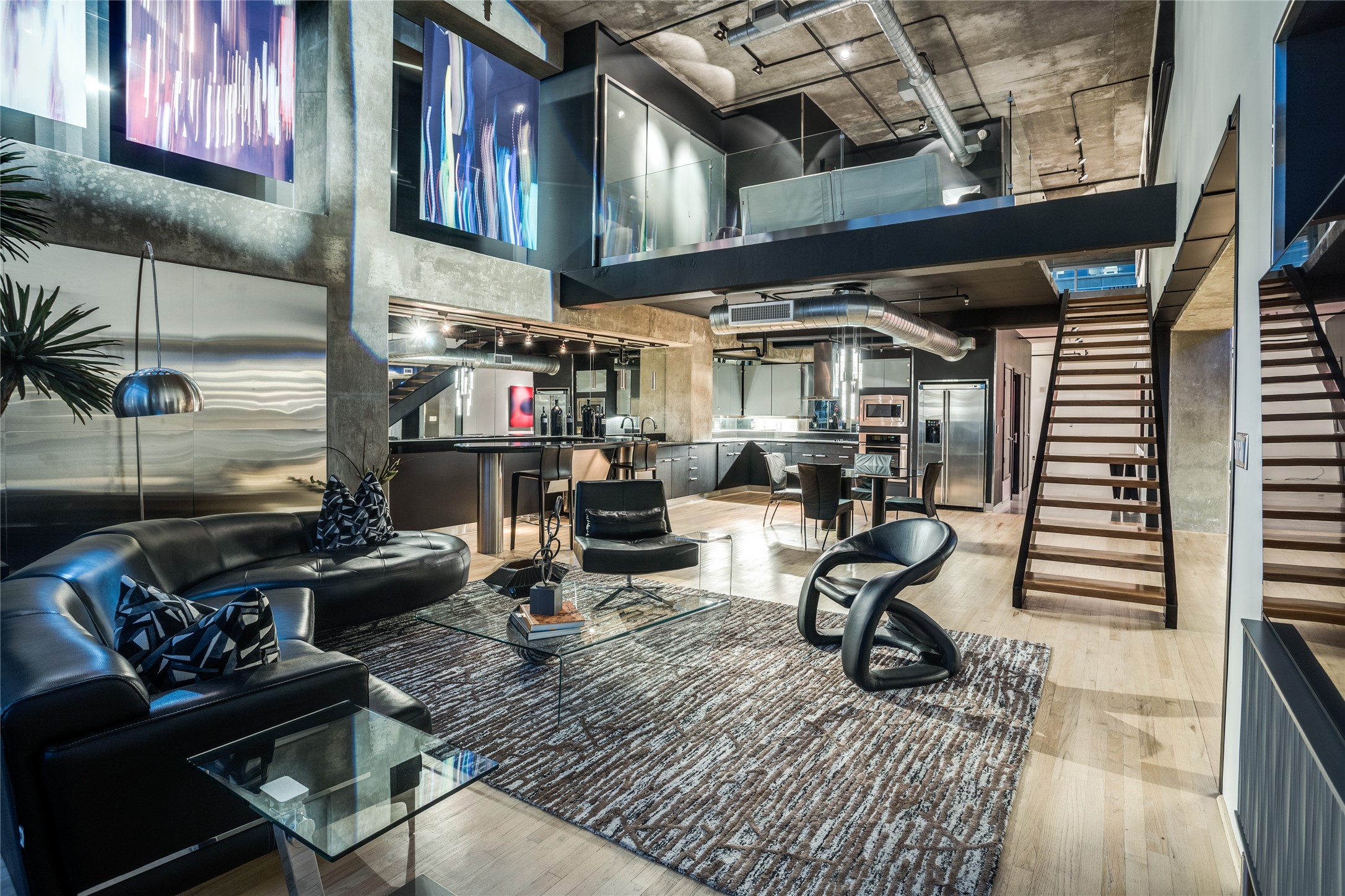 Modern loft living room with black leather seating, an open mezzanine, and a staircase beside the kitchen.