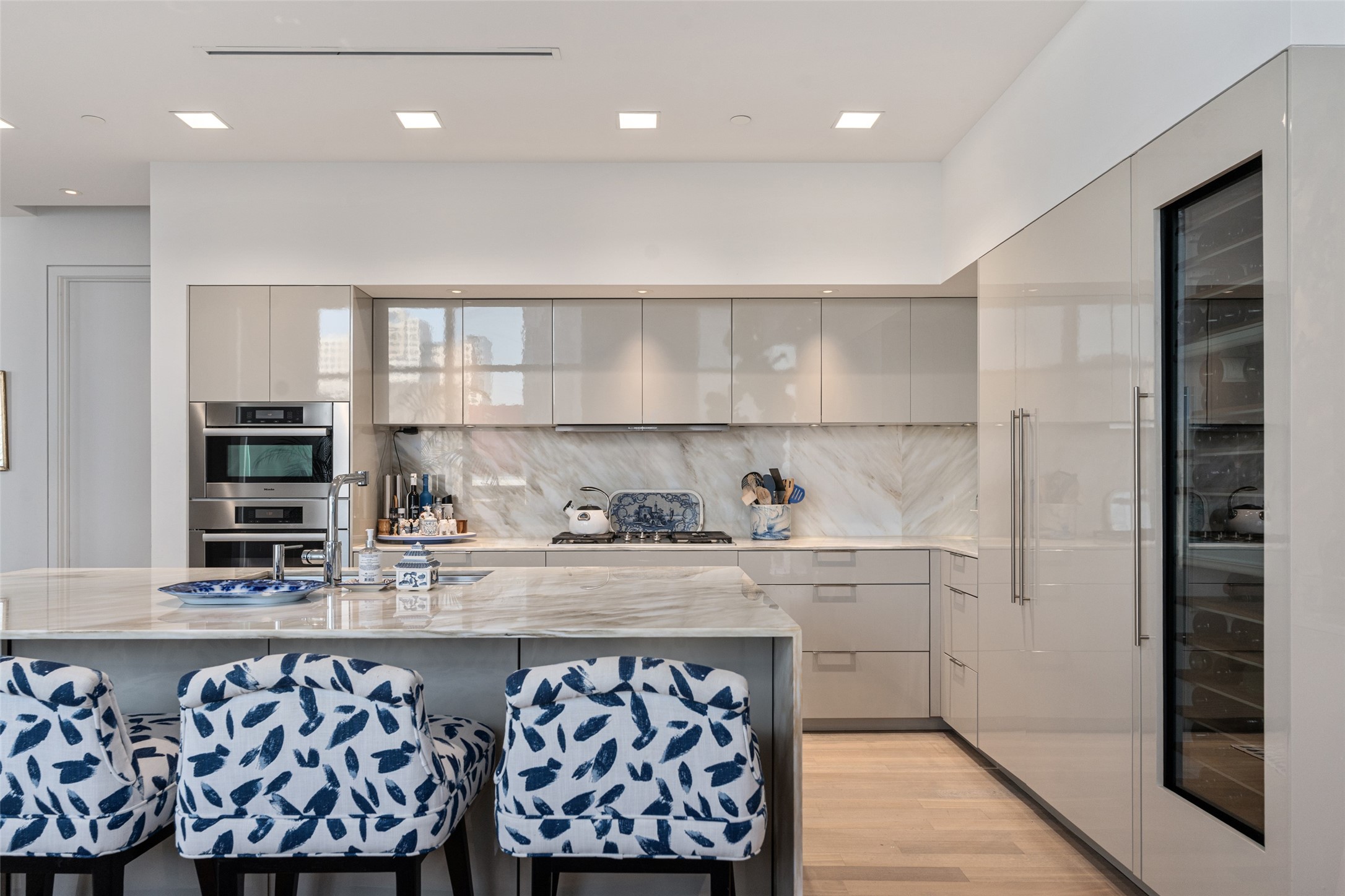 Modern kitchen with a marble island, glossy cabinets, built-in ovens, and navy patterned bar stools.