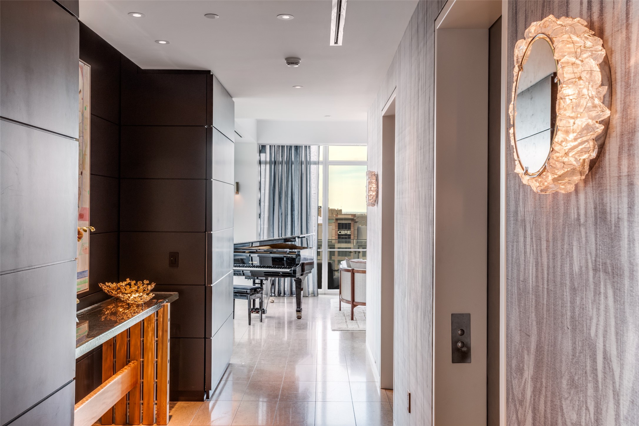 Modern condo hallway with a sculptural illuminated mirror and grand piano by the windows.