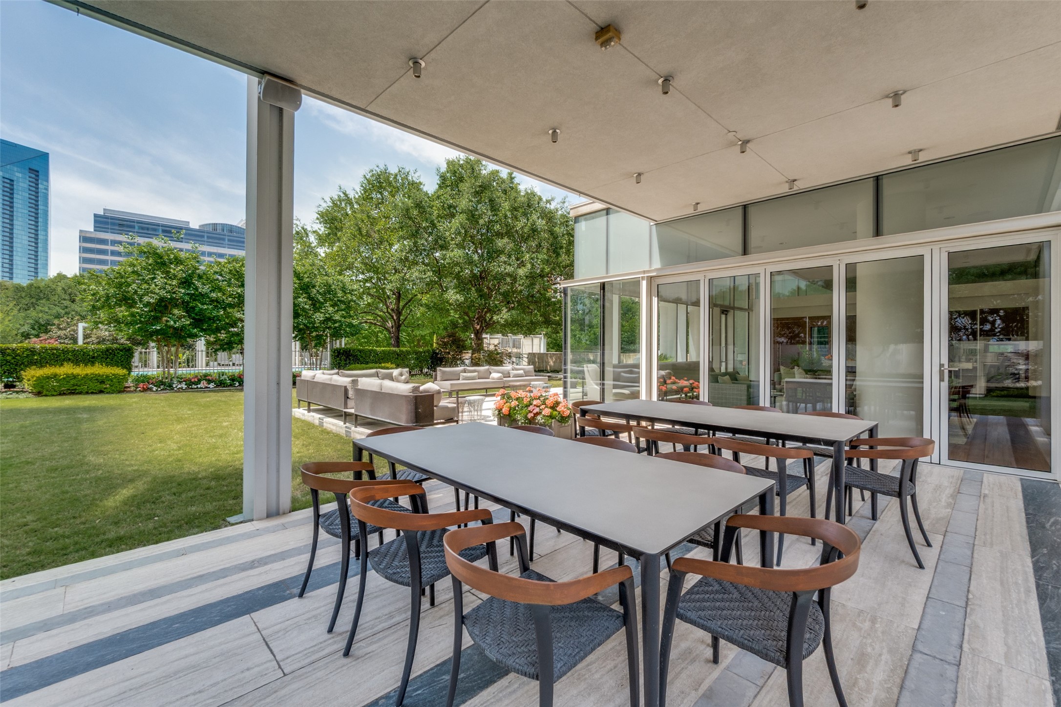 Covered patio with outdoor dining tables, lounge seating, landscaped lawn, and downtown towers beyond.