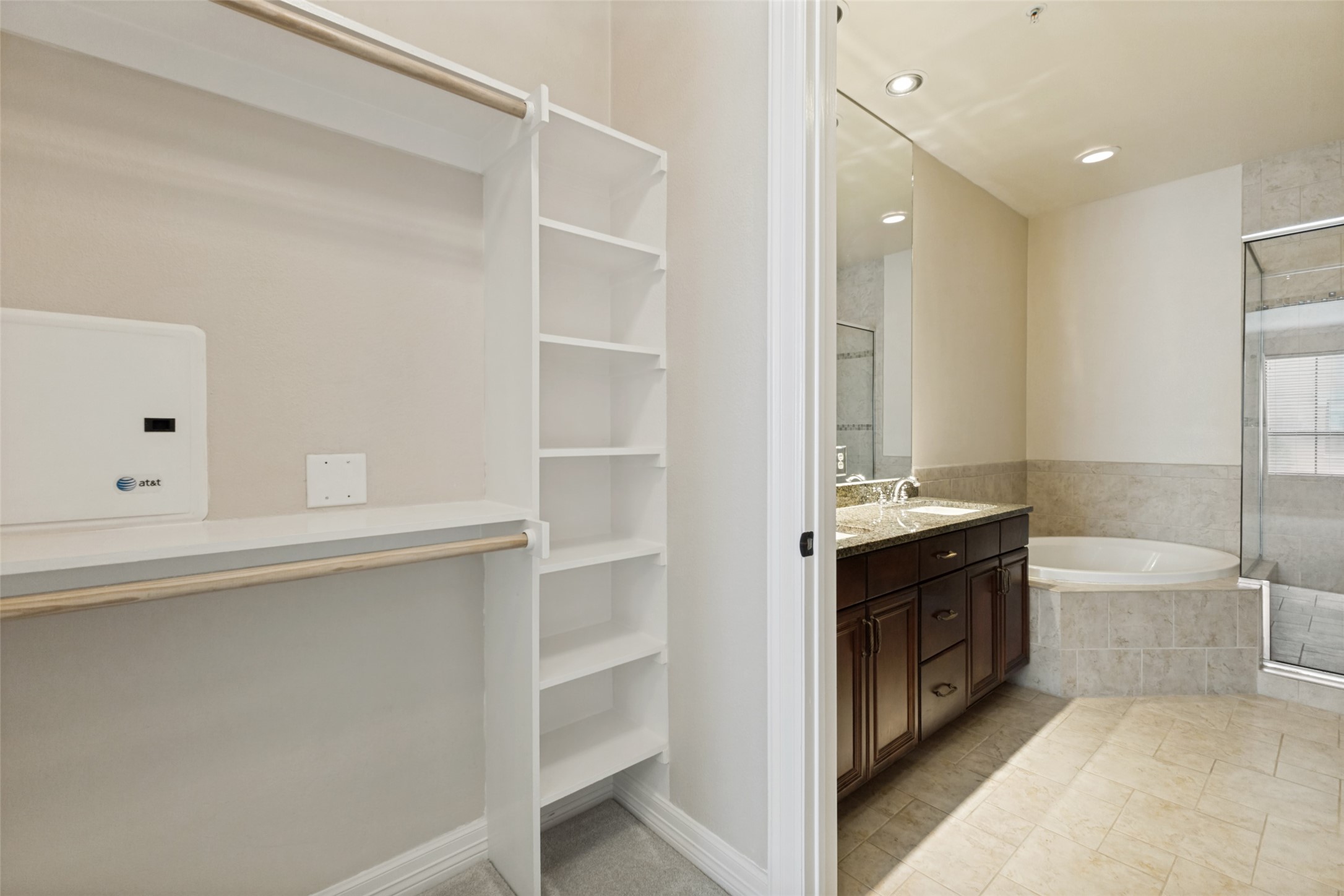 Walk-in closet shelving beside a tiled bathroom with a dark vanity, soaking tub, and glass shower.
