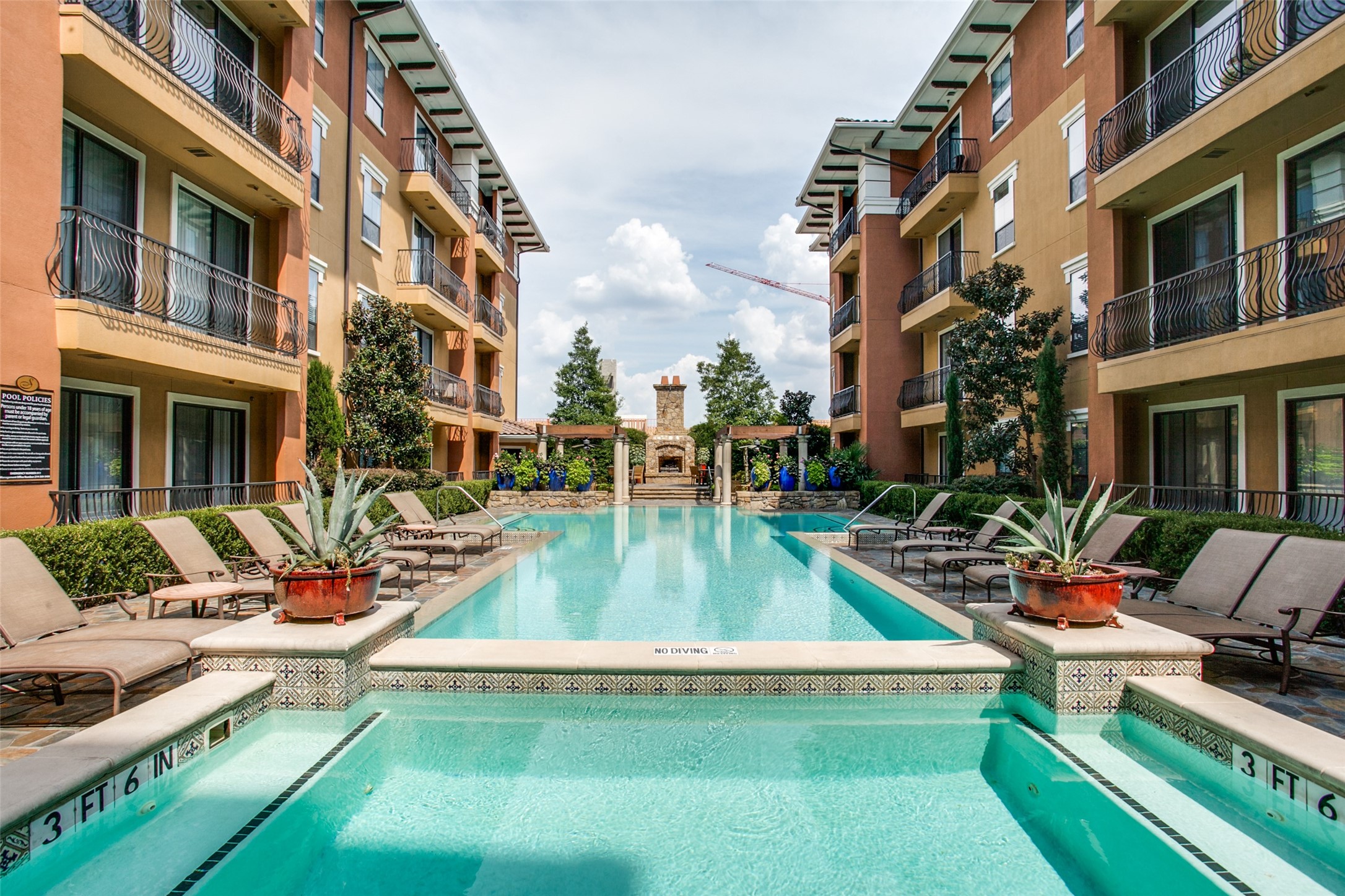 Courtyard swimming pool between apartment buildings, with lounge chairs, potted plants, and a stone fireplace.