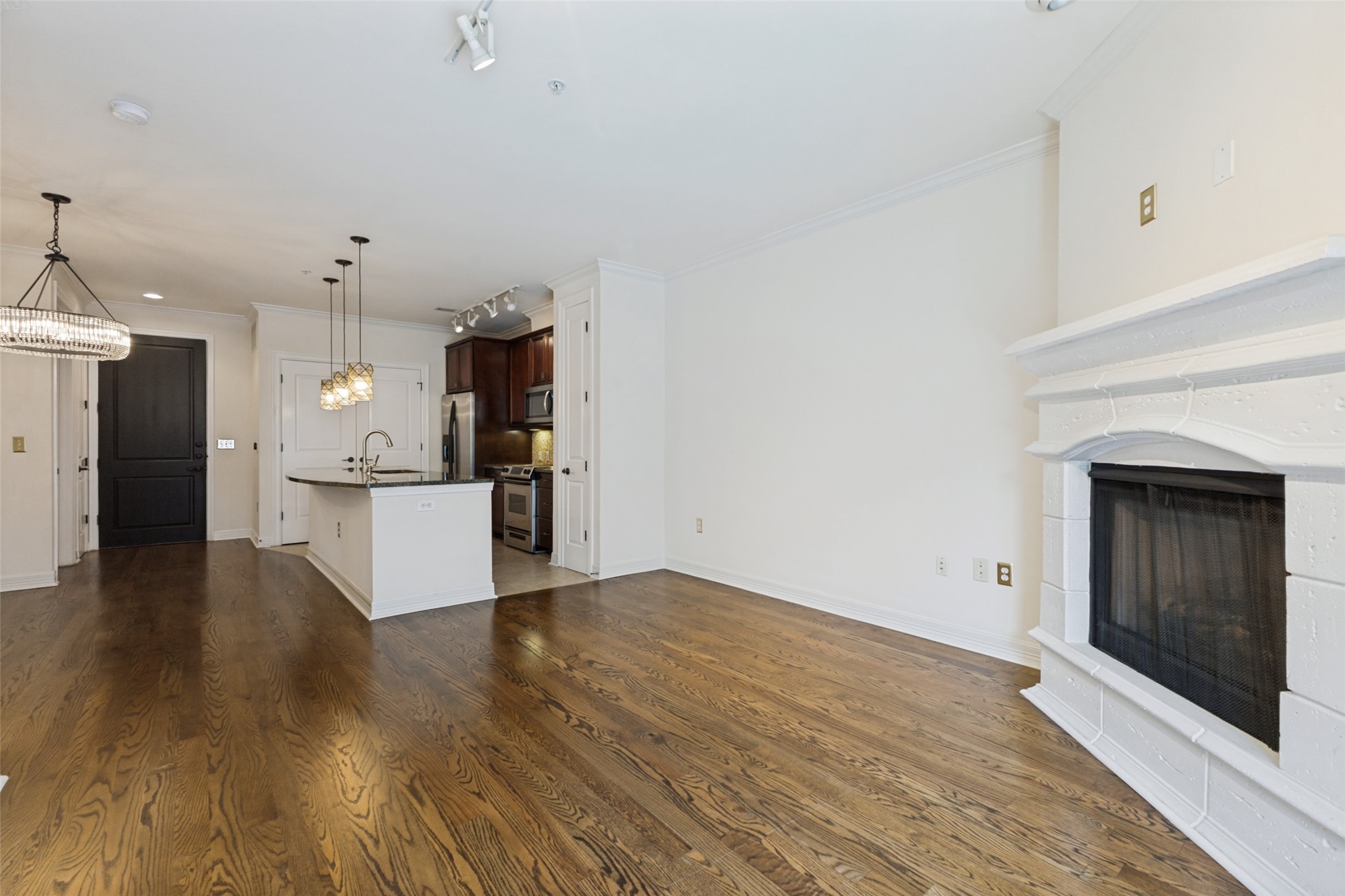 Open-plan living room with hardwood floors and white fireplace beside a kitchen with dark cabinets and island.
