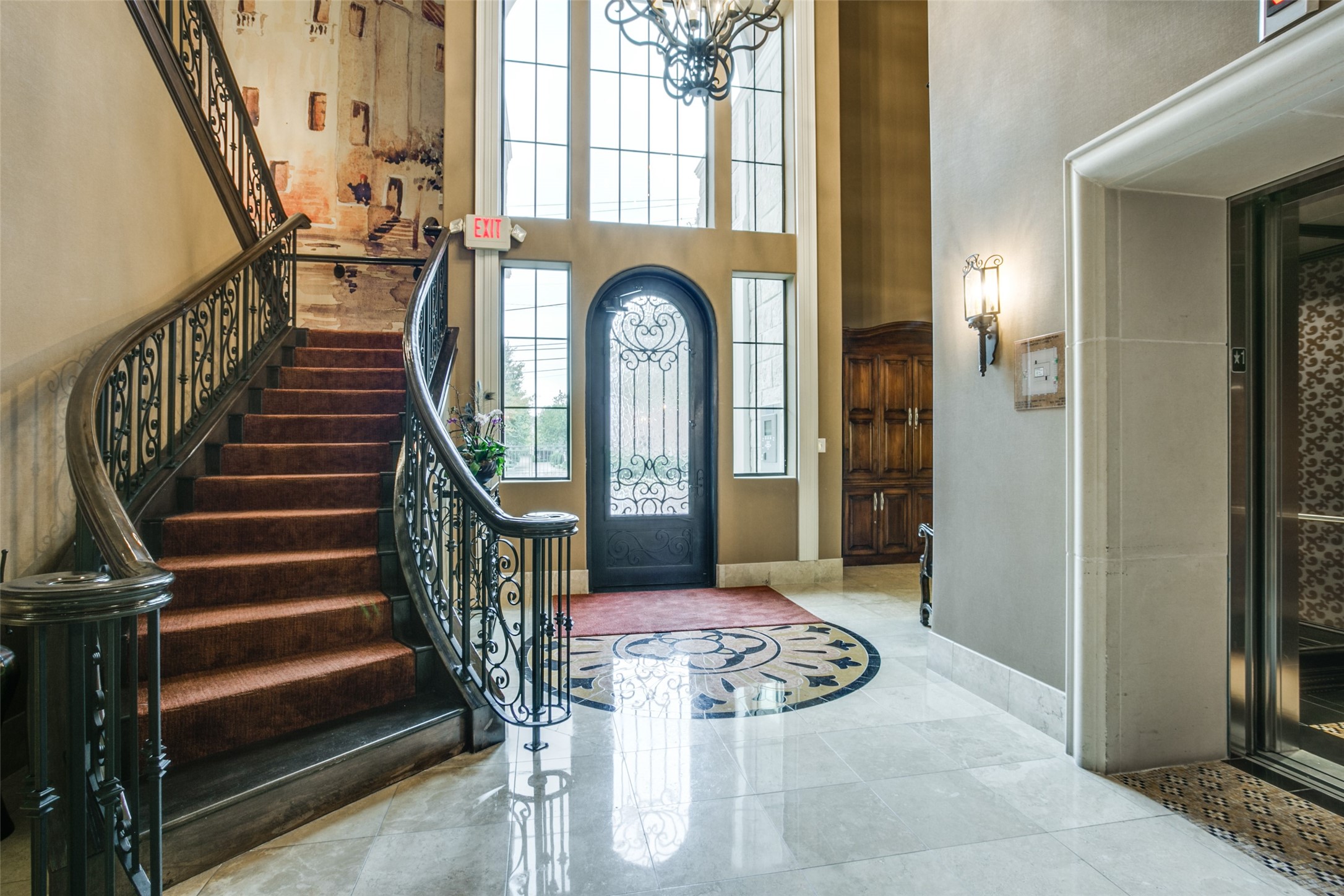 Entry hall with a curved iron-railed staircase, ornate glass front door, circular floor mosaic, and elevator.