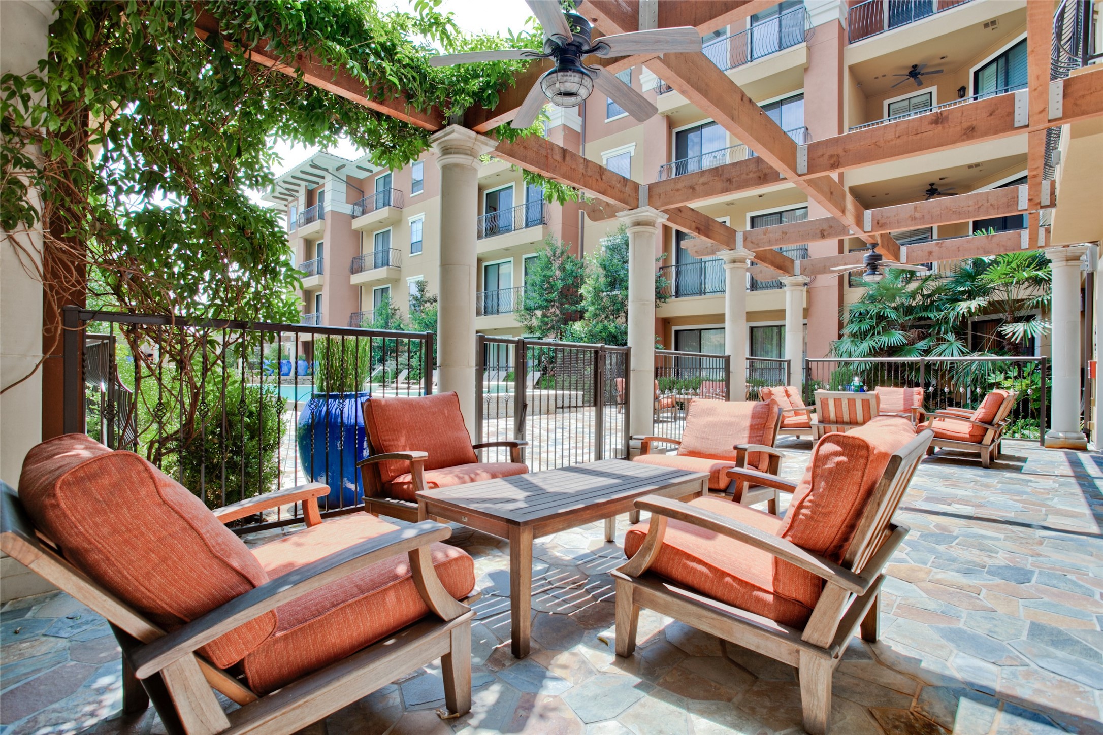 Condominium courtyard patio with cushioned lounge chairs beneath a timber pergola and a pool nearby.