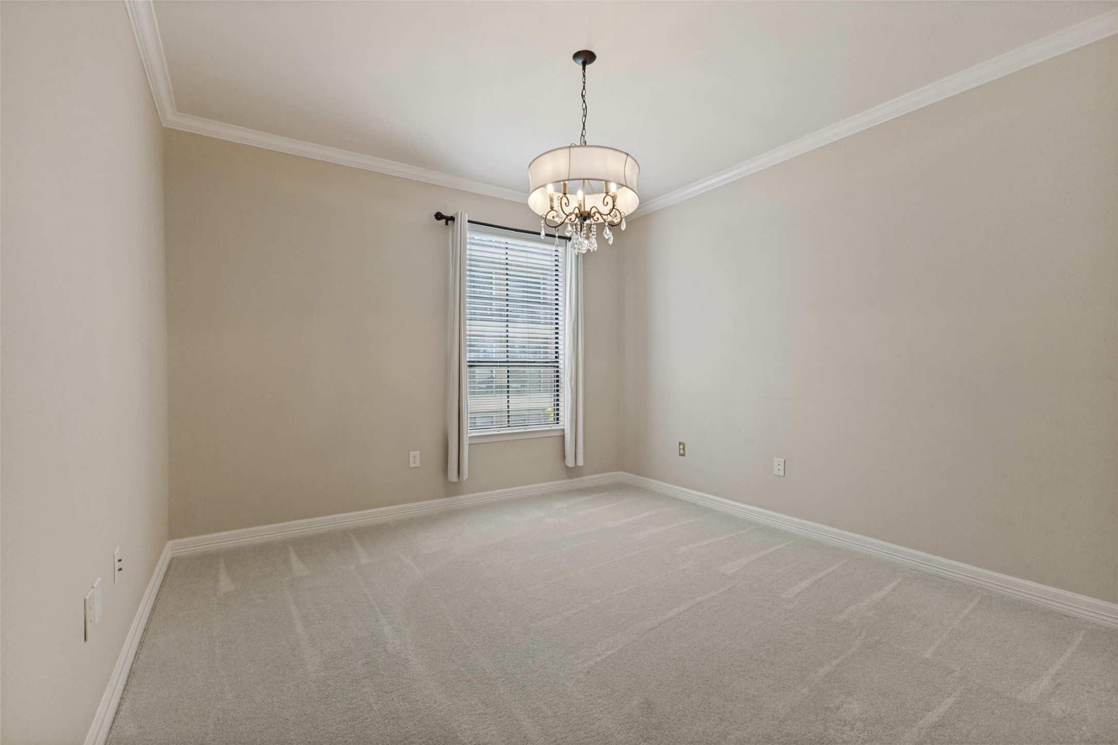 Empty bedroom with a decorative chandelier, carpeted floor, and window with blinds.