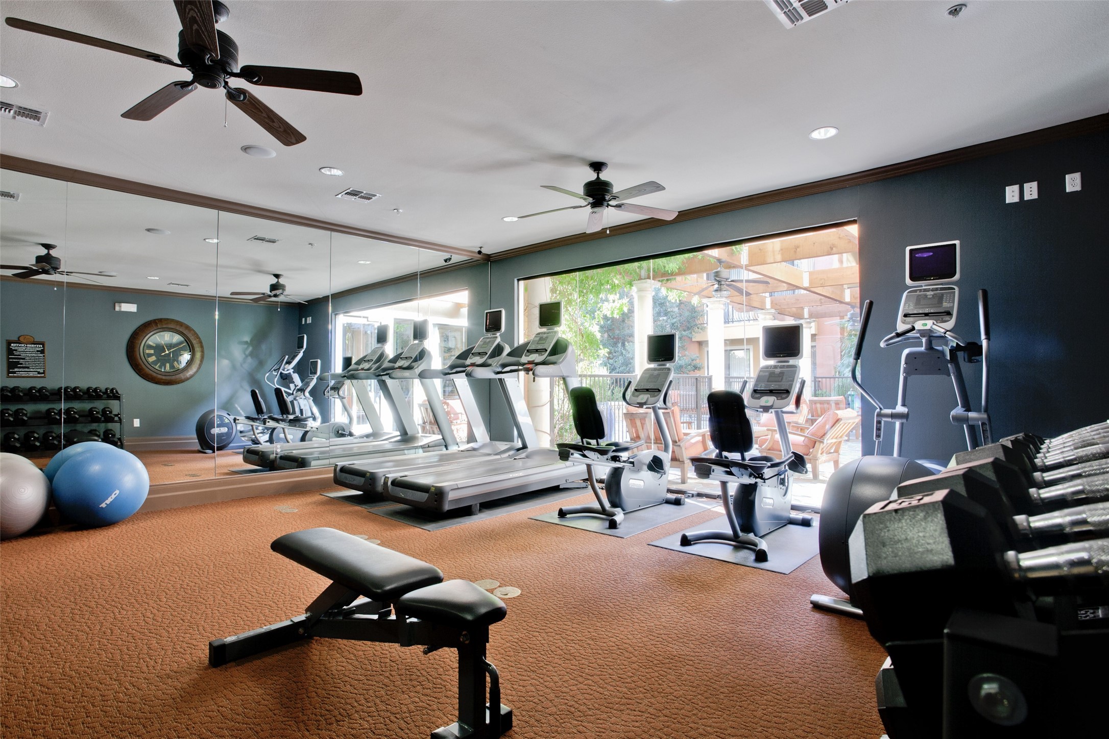 Fitness room with treadmills, bikes, weights, and large windows overlooking a patio.