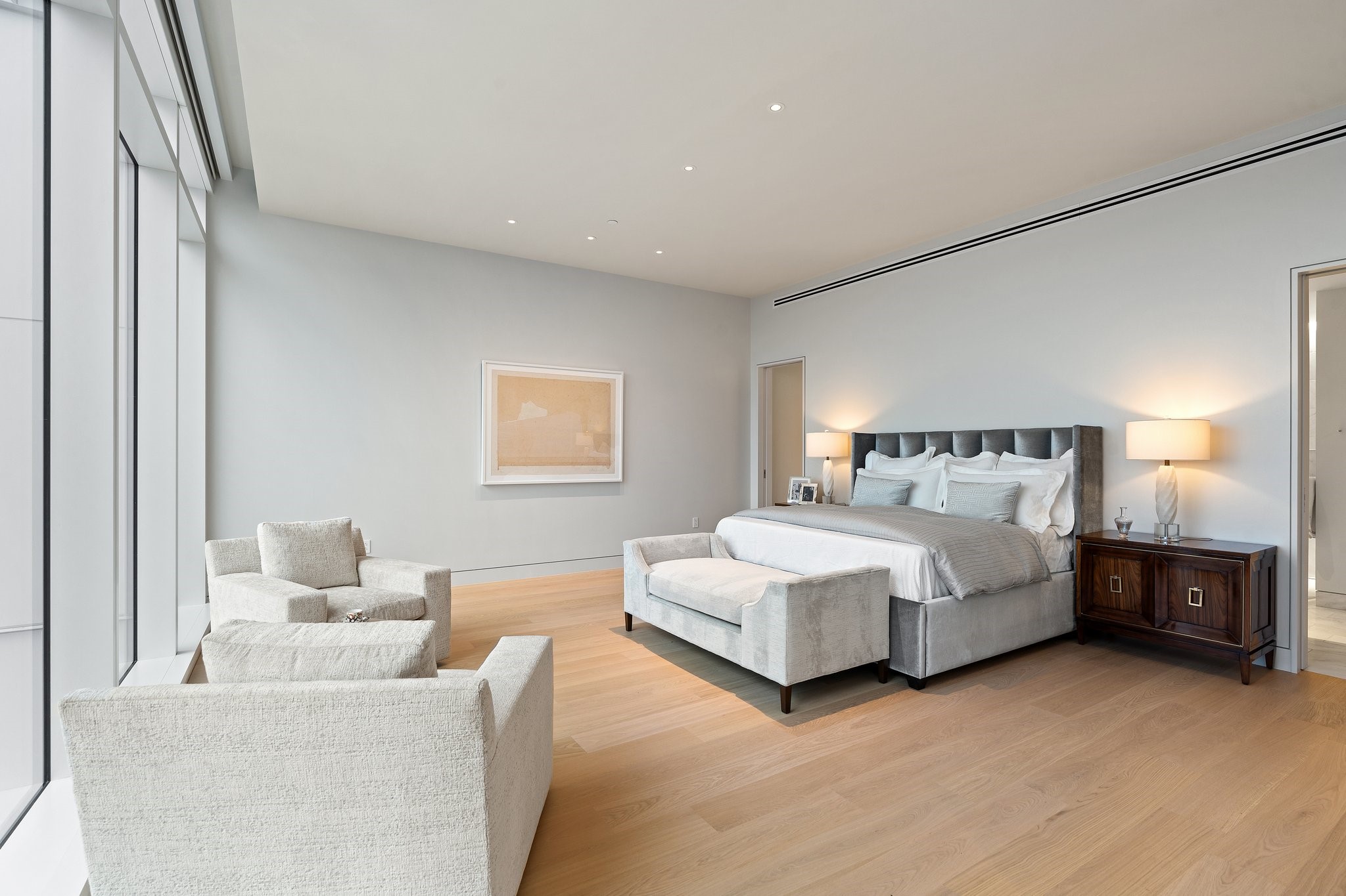 Modern bedroom with a gray upholstered bed, pale seating, and floor-to-ceiling windows.