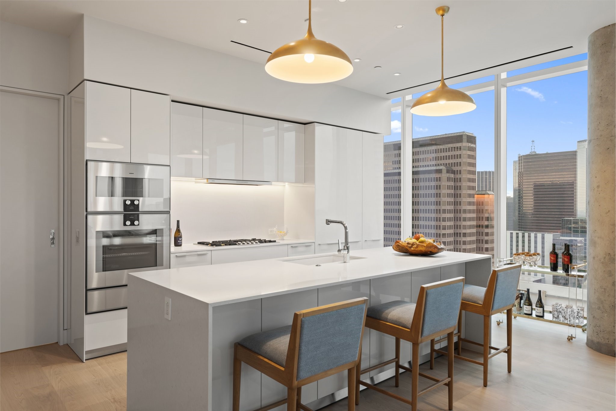 Modern white kitchen with a large island, bar seating, and Dallas skyline views through floor-to-ceiling windows.