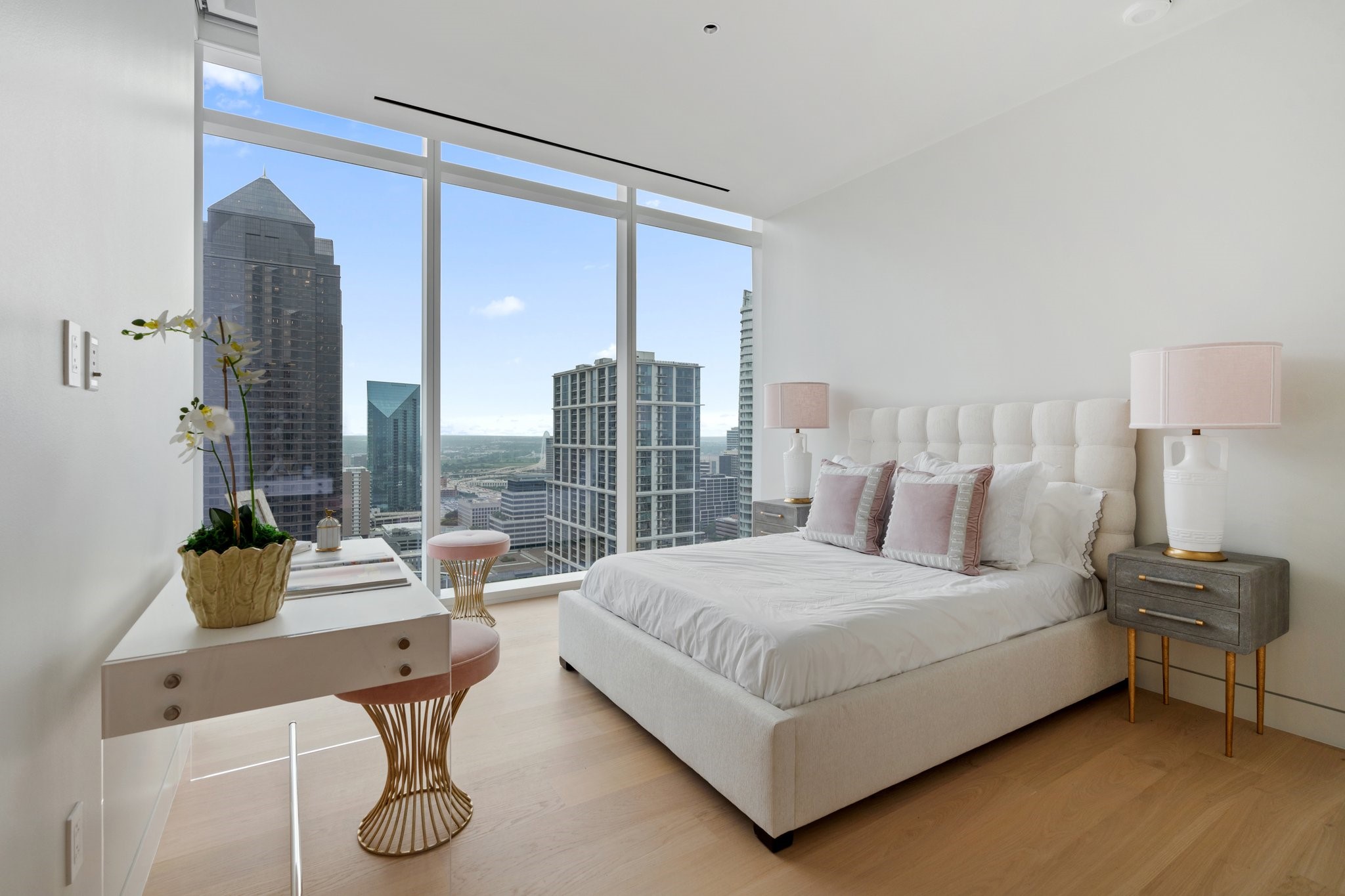 Staged bedroom with a white upholstered bed and floor-to-ceiling windows overlooking the Dallas skyline.