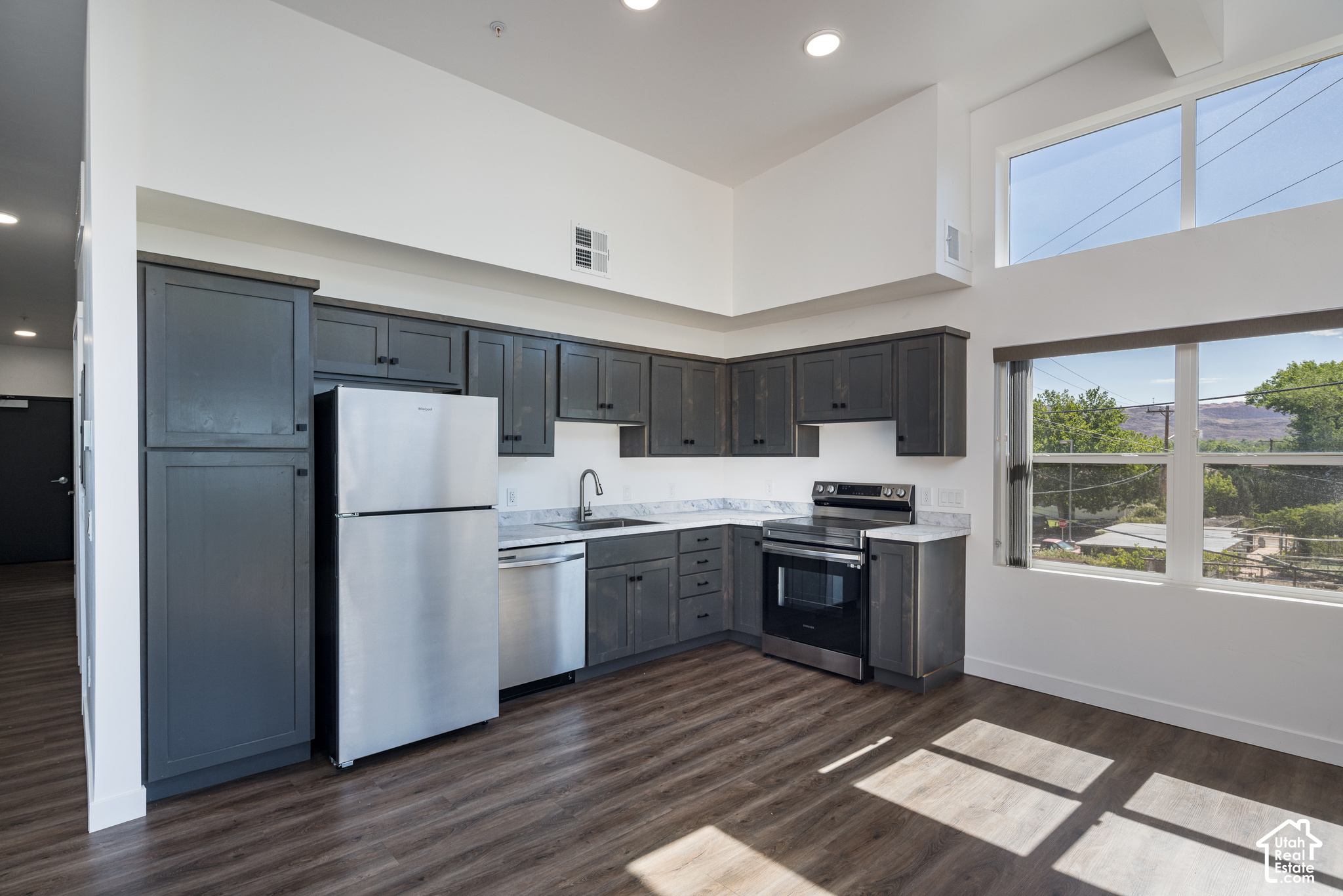 Unfurnished kitchen with dark gray cabinets, stainless steel appliances, and tall windows overlooking trees and rocky terrain.