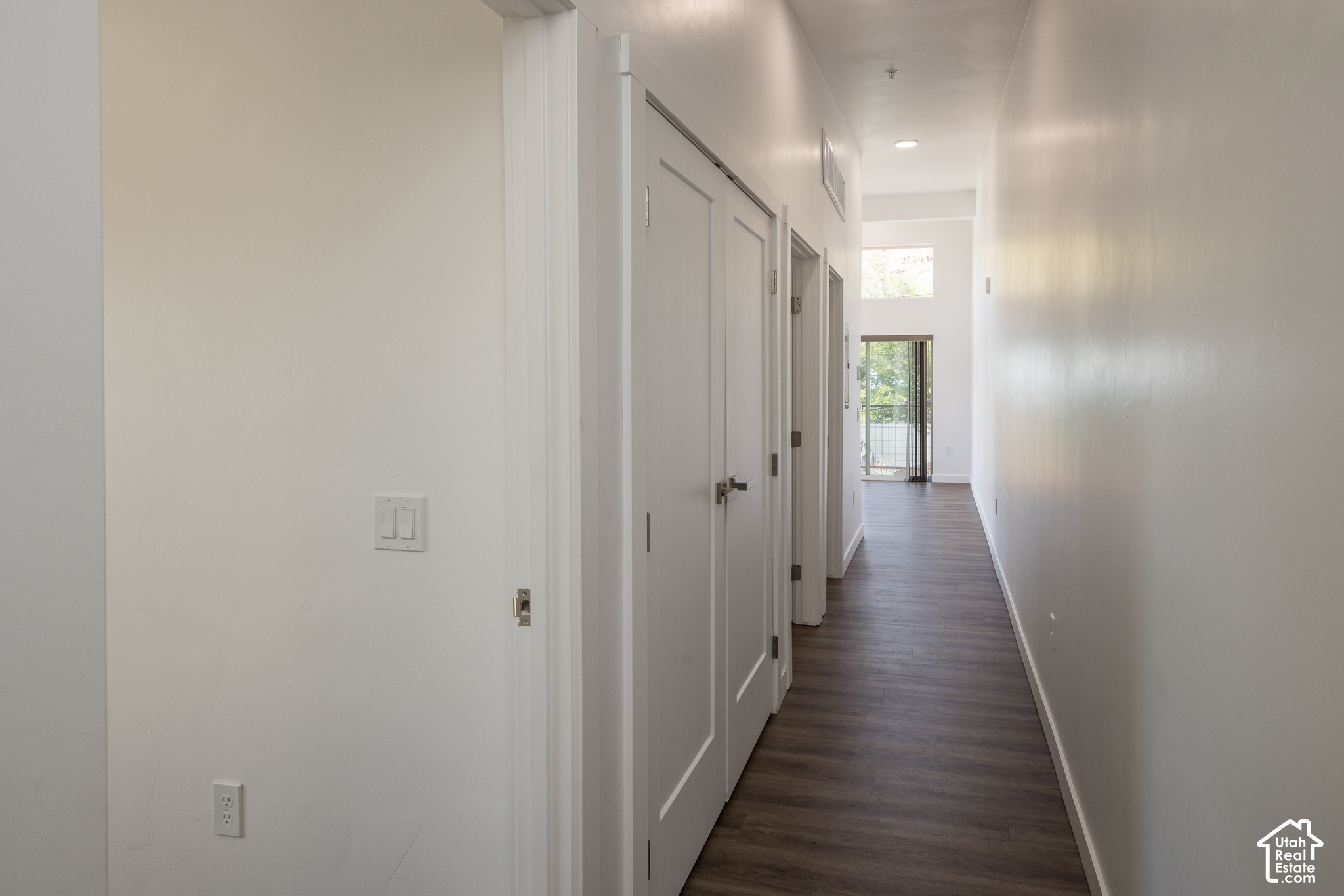 Empty hallway with dark flooring, white doors, and a glass balcony door at the far end.