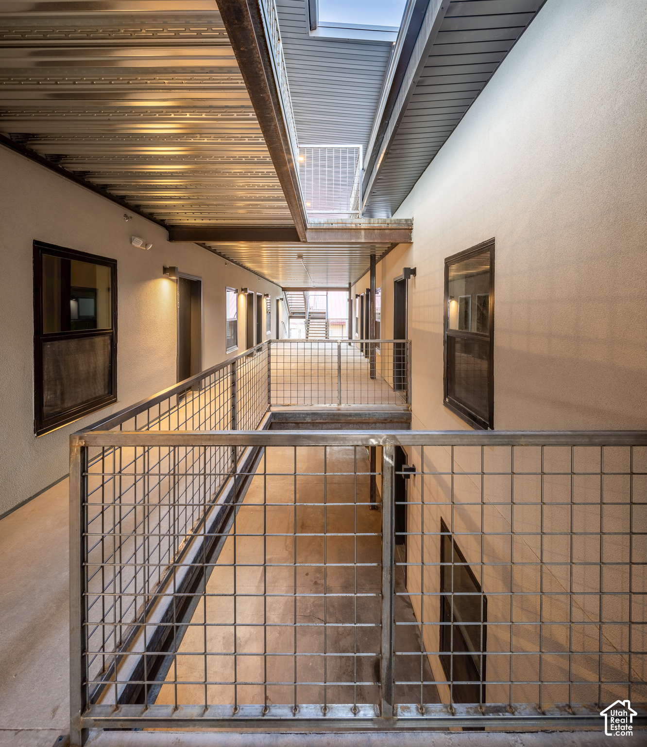Apartment corridor with metal grid railings overlooking a lower-level walkway beneath an exposed metal ceiling.