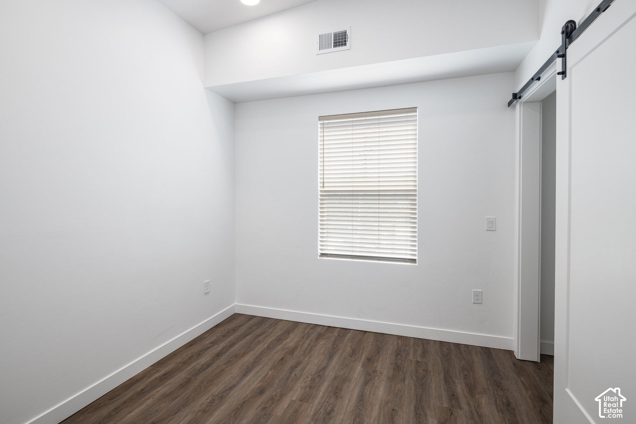 Empty room with dark wood-look flooring, a window with blinds, and a black sliding barn door.