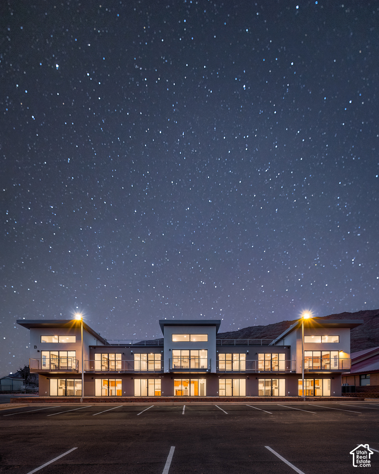 Modern two-story condominium building glowing beneath a star-filled night sky, with balconies and a front parking lot.