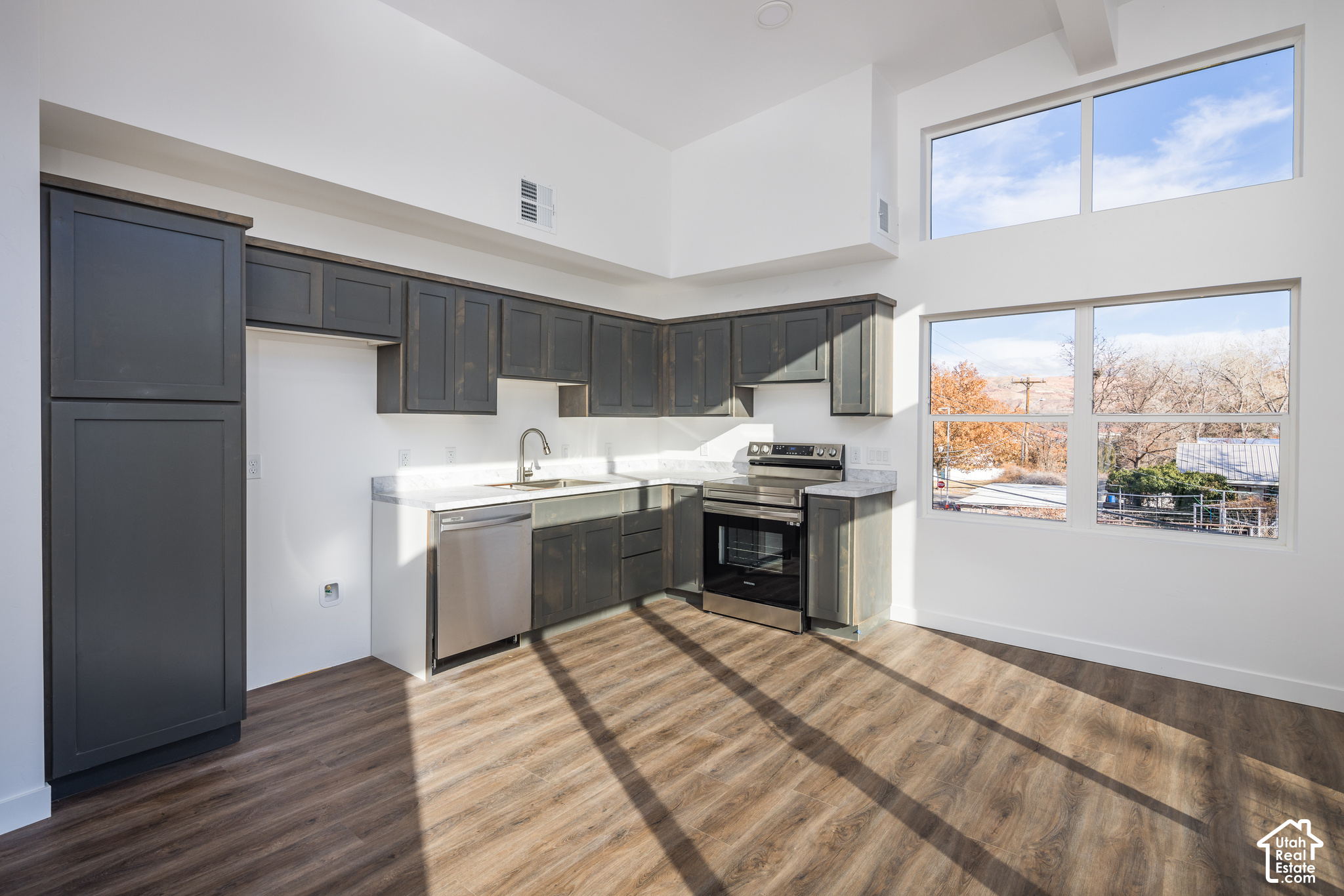 Bright vacant kitchen with charcoal cabinets, stainless appliances, and tall sunlit windows.