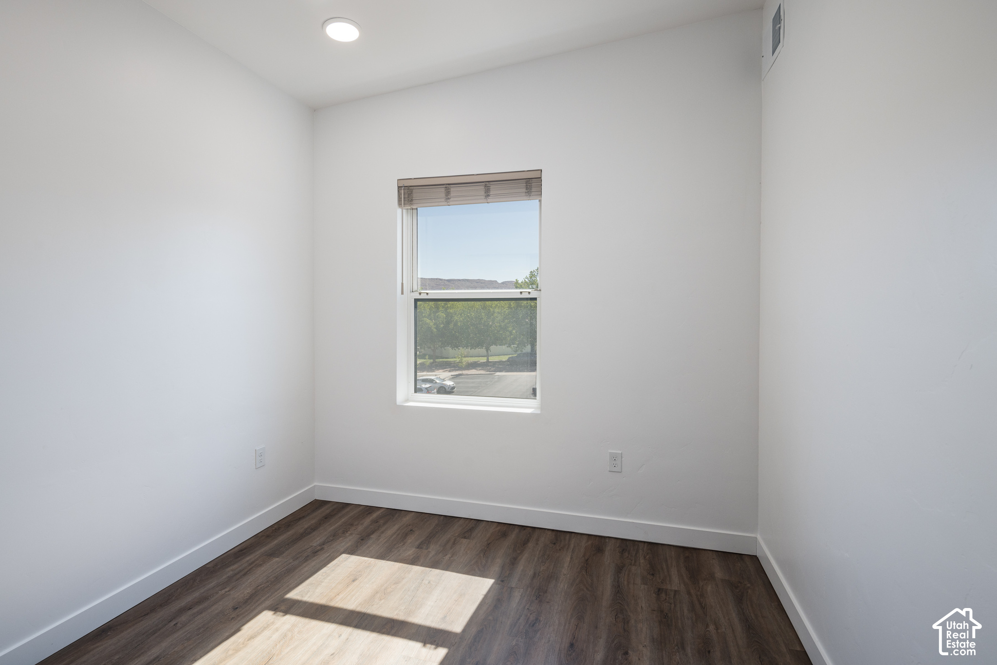 Empty bedroom with dark wood-look flooring and a sunlit window overlooking trees.