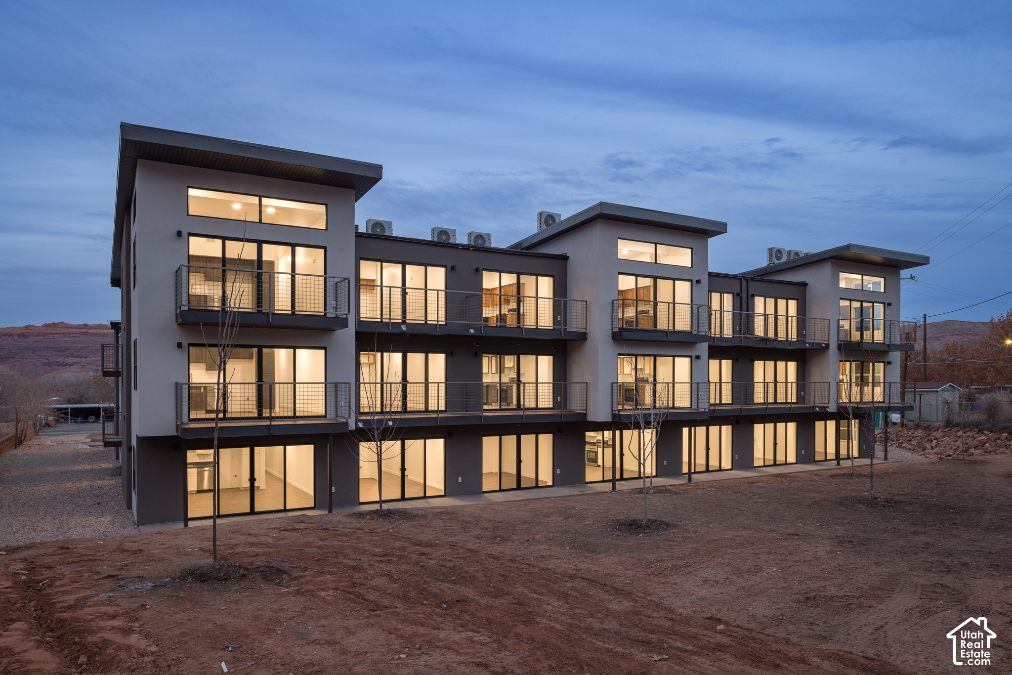 Modern residential building with illuminated windows and balconies at dusk, set against low rocky hills.