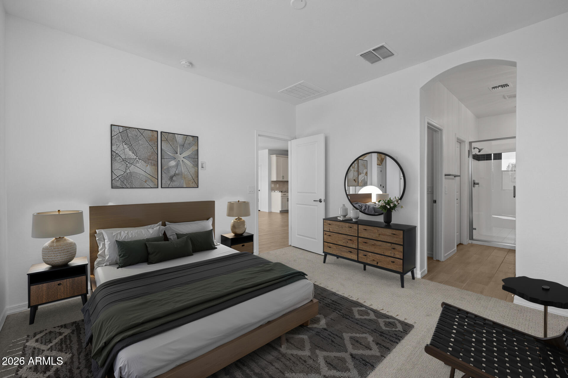 Staged bedroom with gray and green bedding, a wood dresser and round mirror, and an adjoining bathroom.