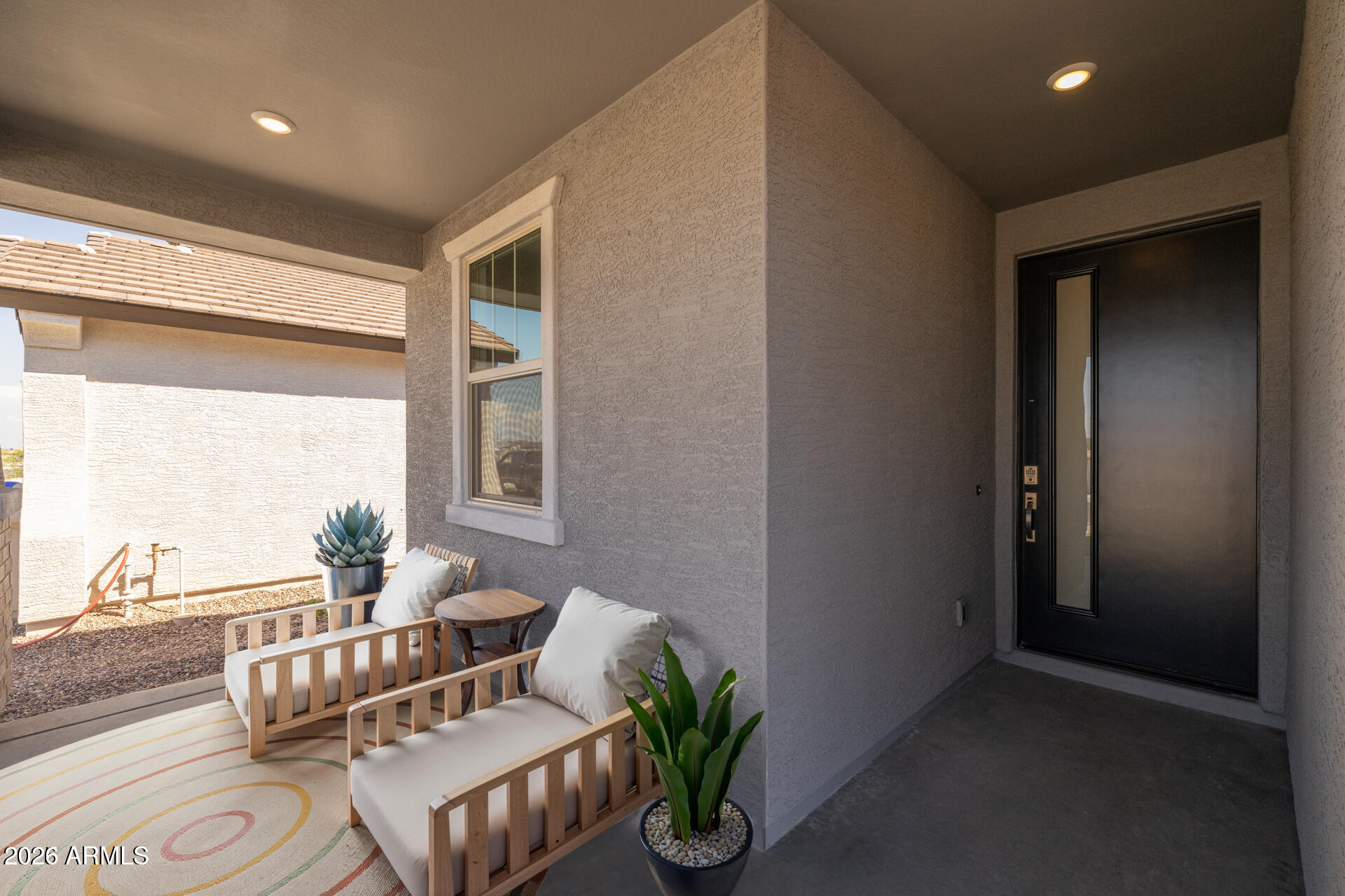 Covered front porch with a dark entry door, cushioned wooden chairs, potted plants, and a patterned rug.