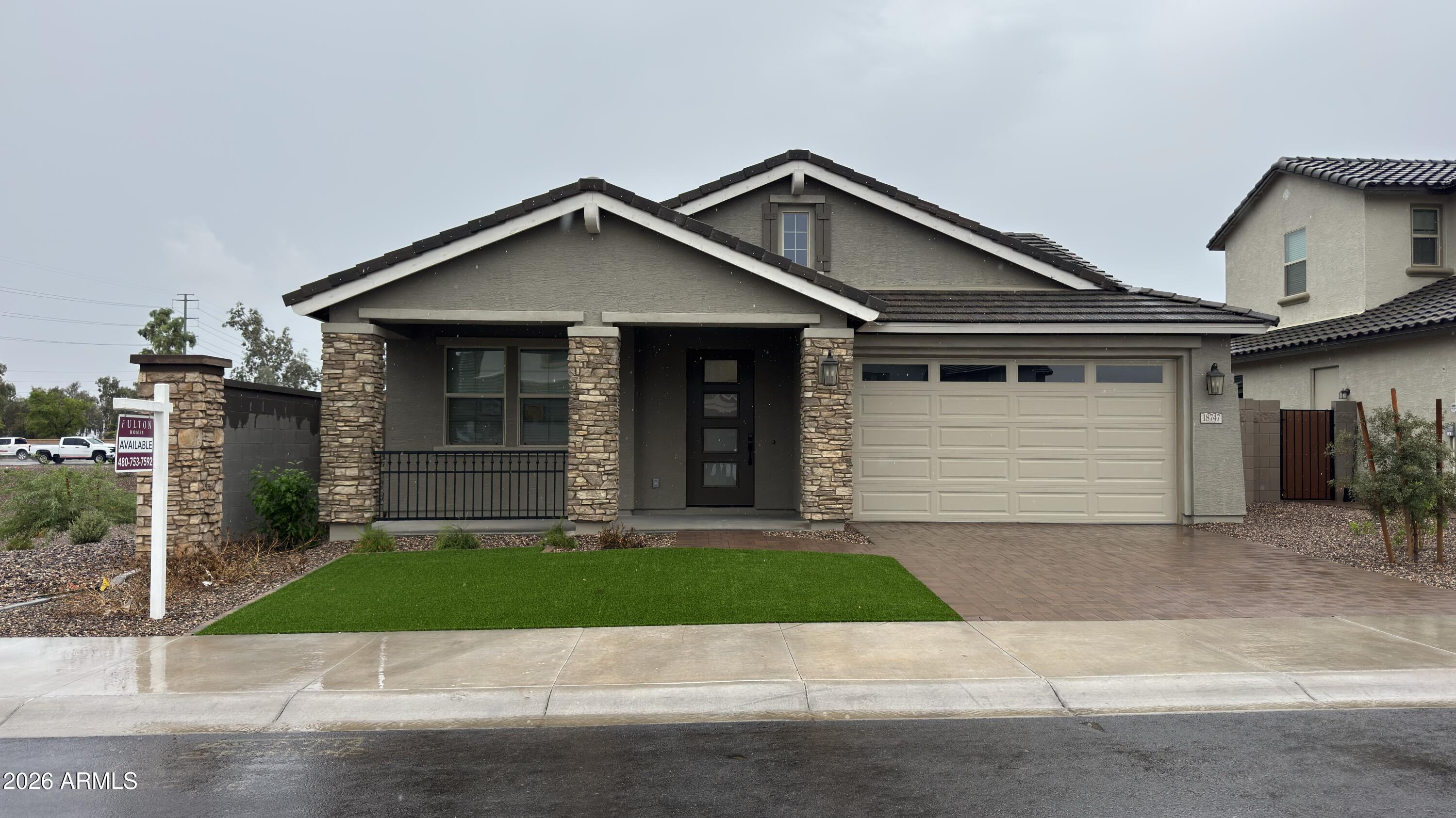 Gray stucco home with stone porch columns, dark gabled roof, two-car garage, and a small front lawn.
