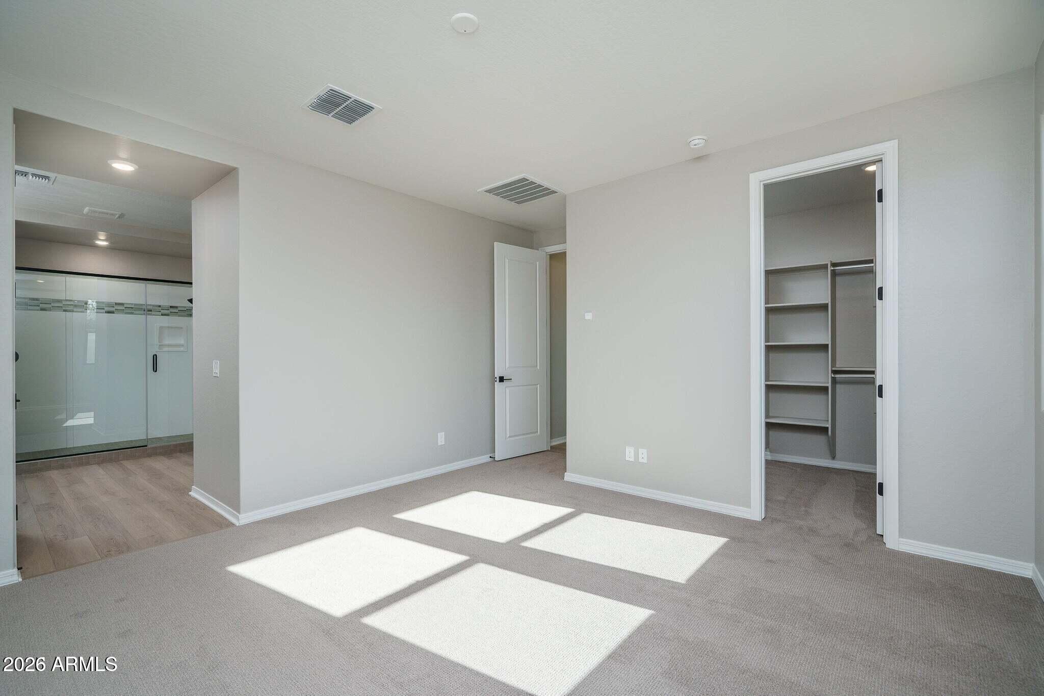 Unfurnished bedroom with a walk-in closet, adjoining glass shower, and sunlight across the carpet.