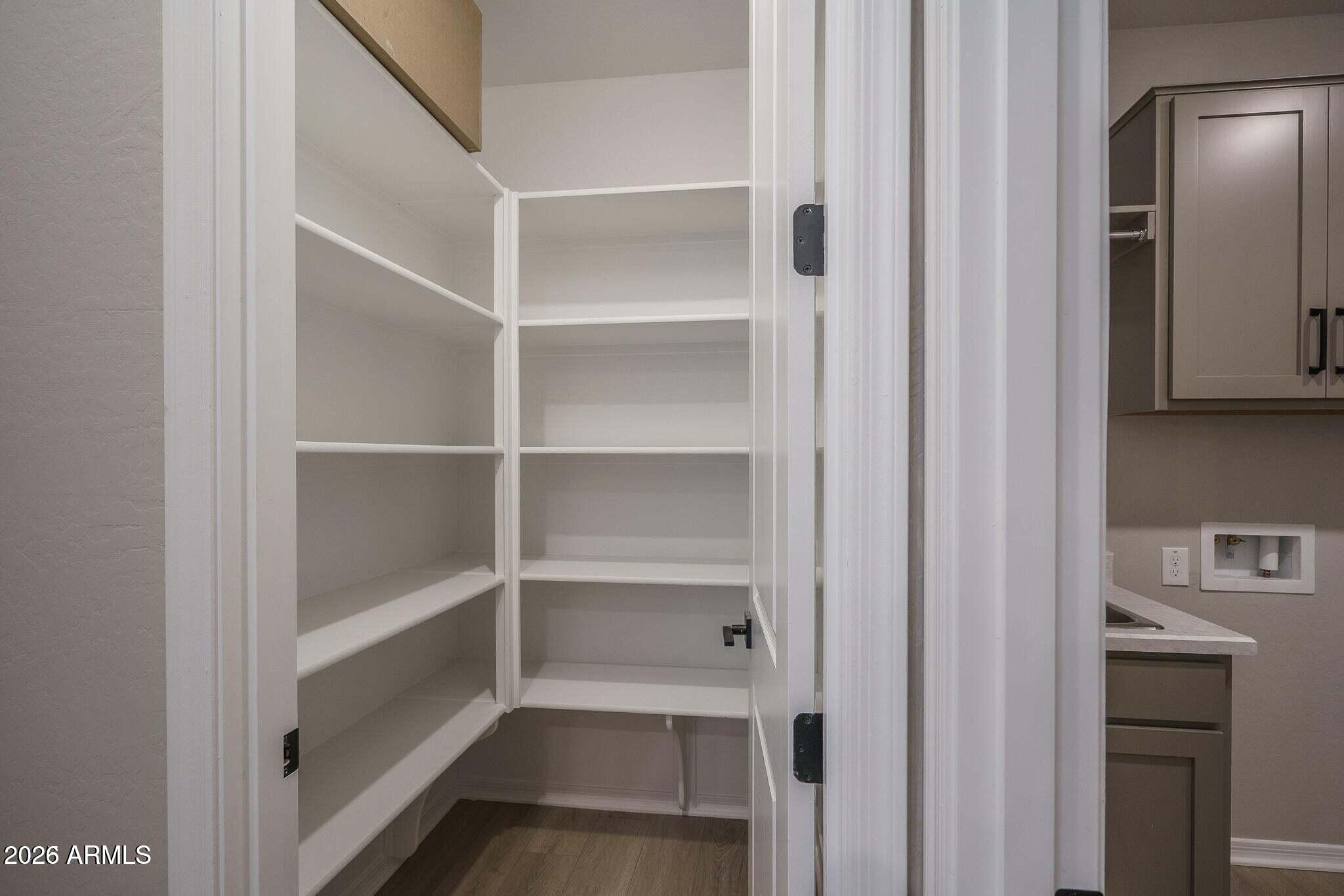 Walk-in pantry with empty white shelves and wood flooring beside gray kitchen cabinets.