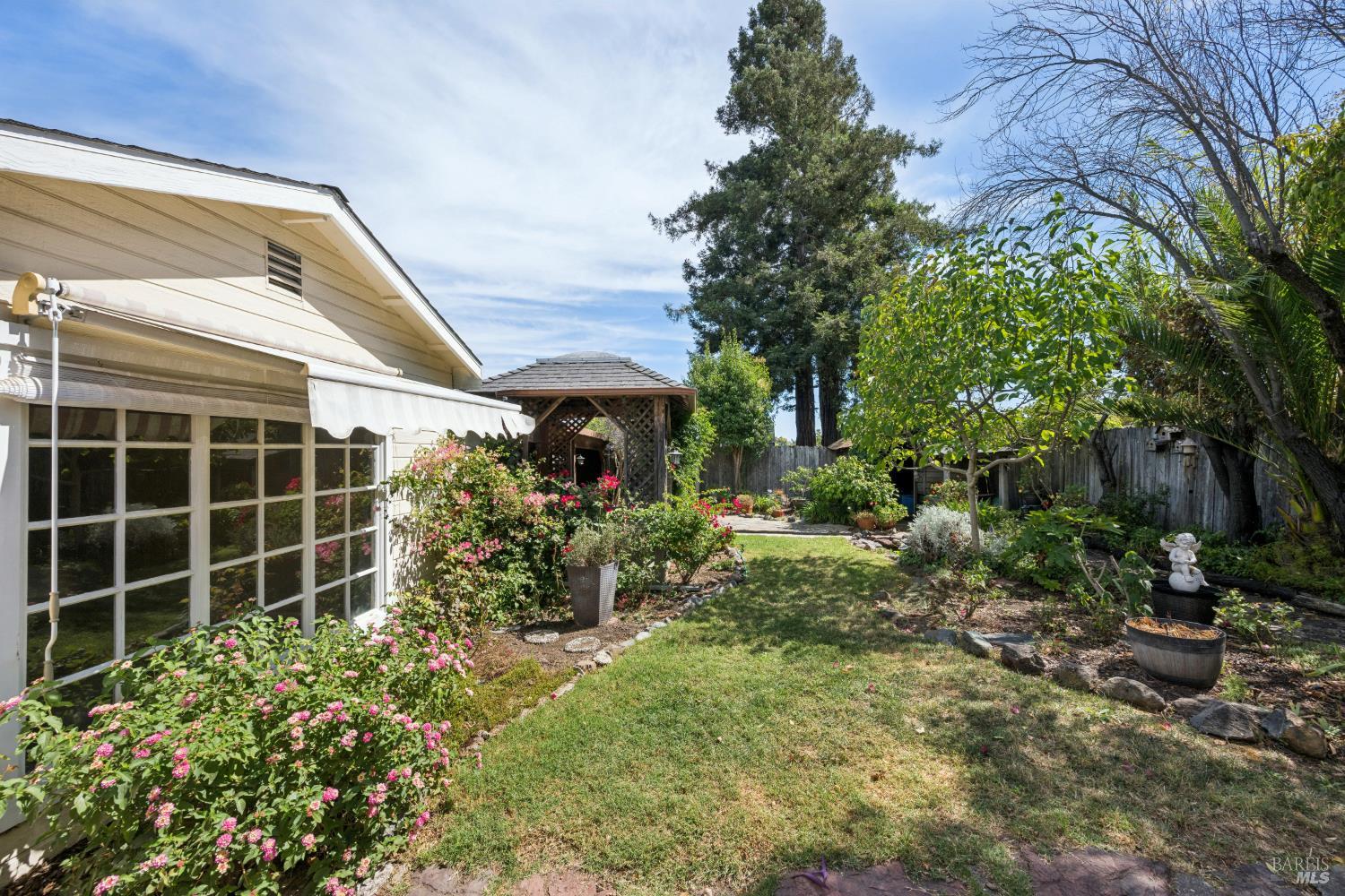 Landscaped backyard with pink roses, a lawn, mature trees, and a wooden gazebo.