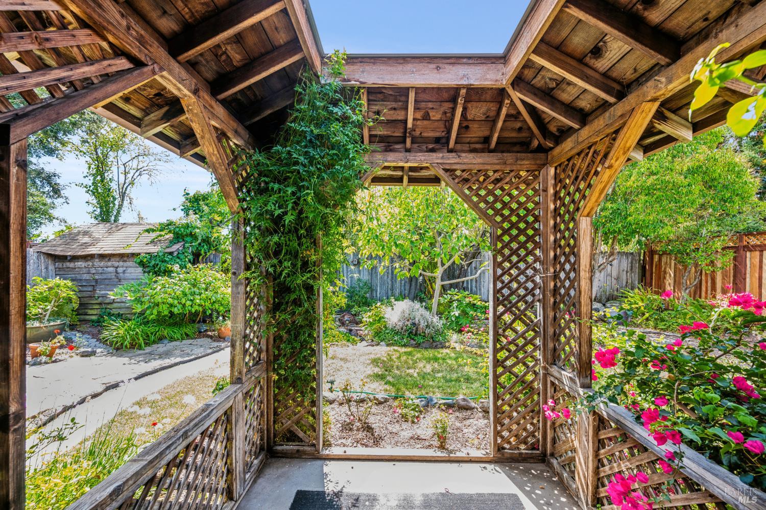 Covered wooden patio with lattice sides overlooking a garden of flowering plants and shrubs.