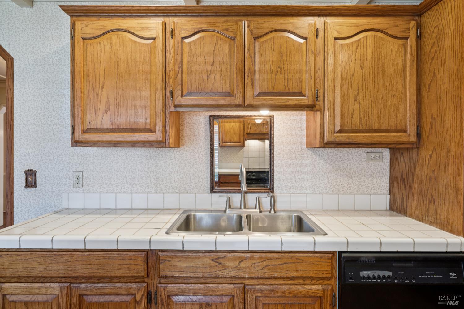 Kitchen sink beneath honey-toned wood cabinets, with a tiled countertop and black dishwasher.