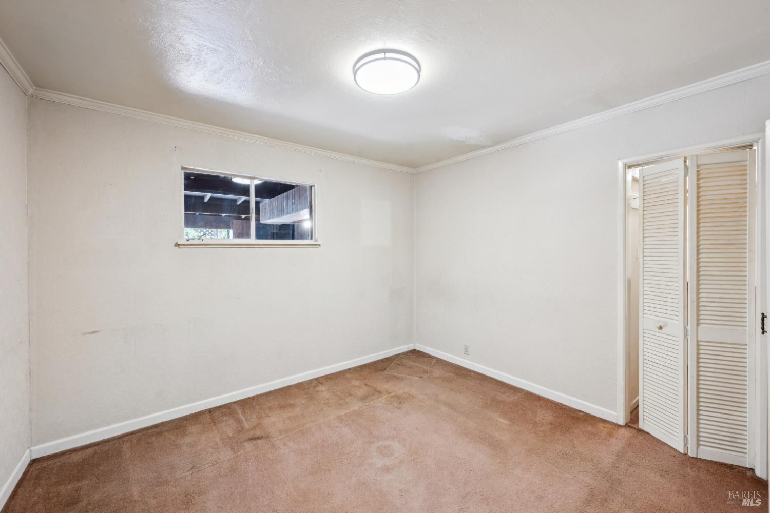 Empty bedroom with tan carpet, a small window, and louvered closet doors.