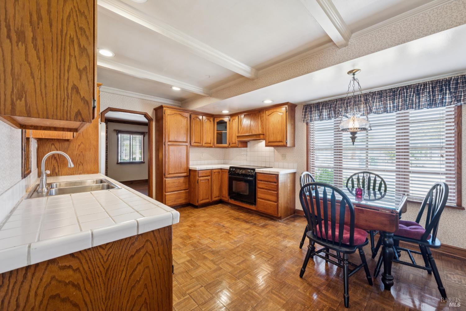 Kitchen with honey-toned wood cabinets, white tile counters, and an adjoining dining area by a large window.