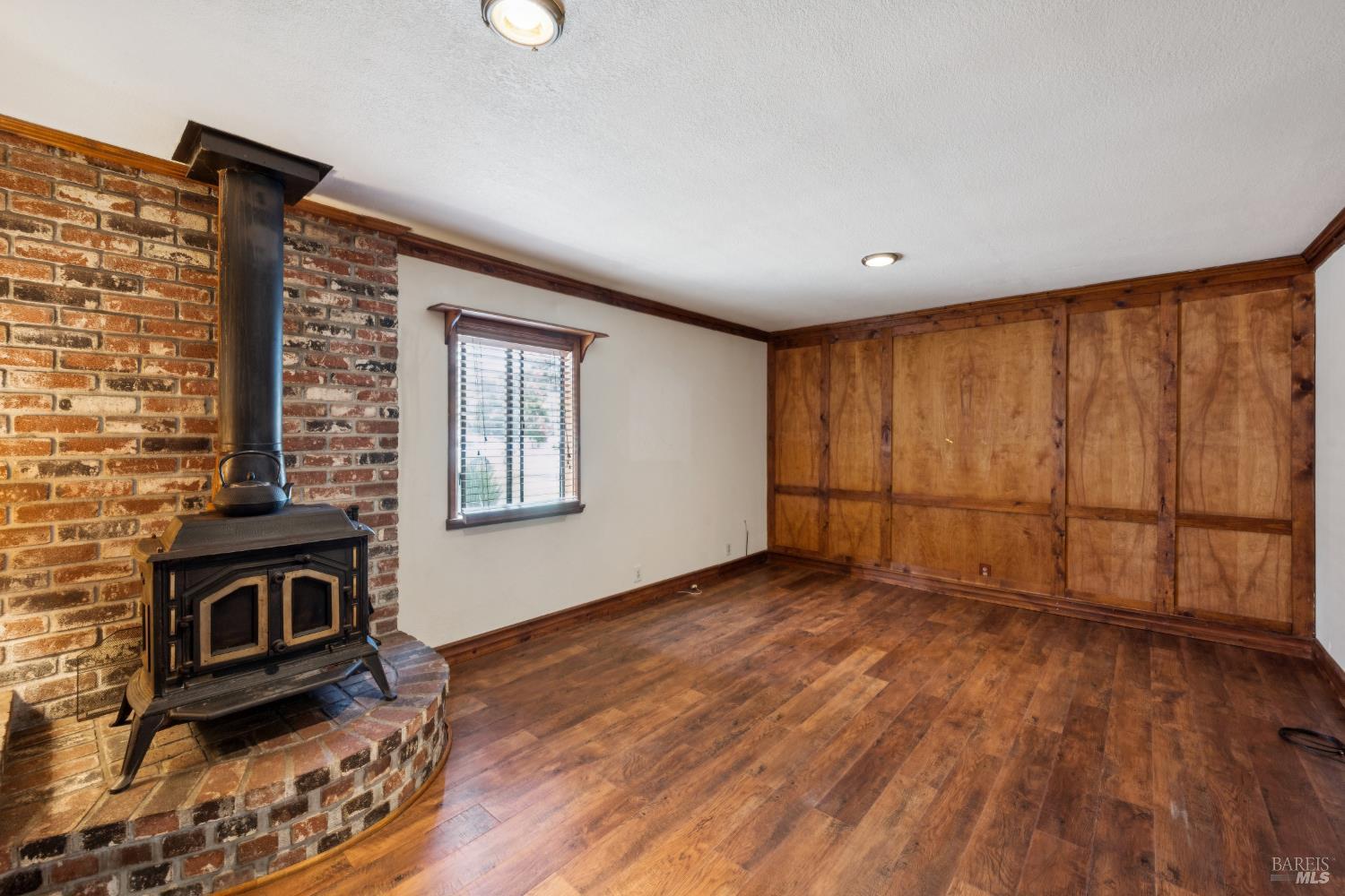 Unfurnished room with a freestanding wood stove, brick hearth, wood floors, and paneled walls.