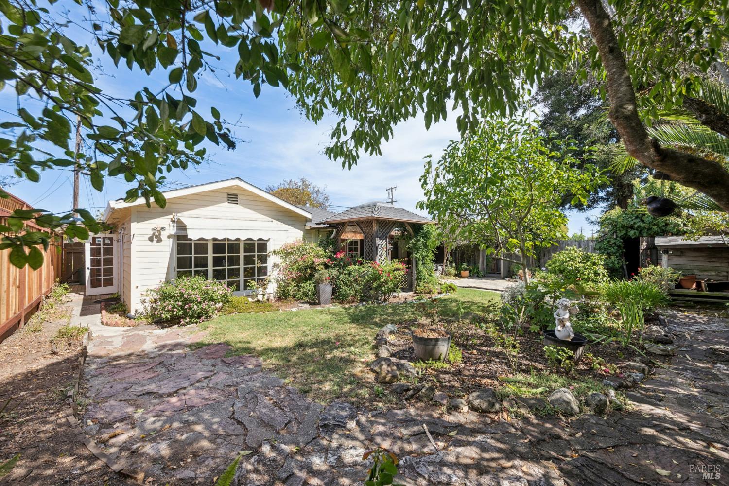 Backyard with a stone patio, flowering garden, and gazebo beside a single-story home.
