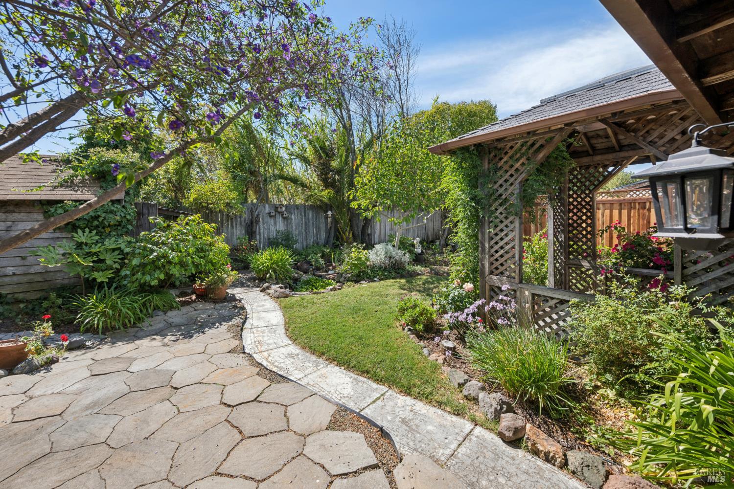 Landscaped backyard with a stone patio, curved walkway, flowering plants, and wooden pergola.