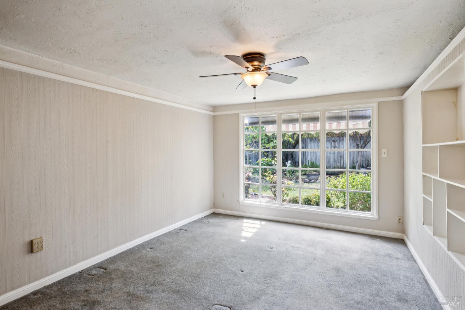 Empty carpeted room with built-in shelving, ceiling fan, and a wide window overlooking a garden.
