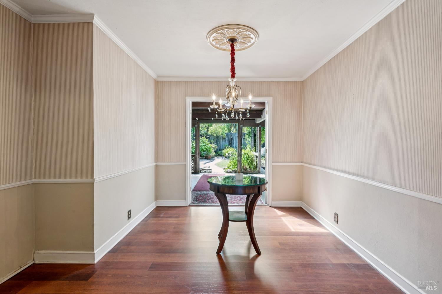 Dining room with a glass-topped table and chandelier, opening onto a leafy garden.
