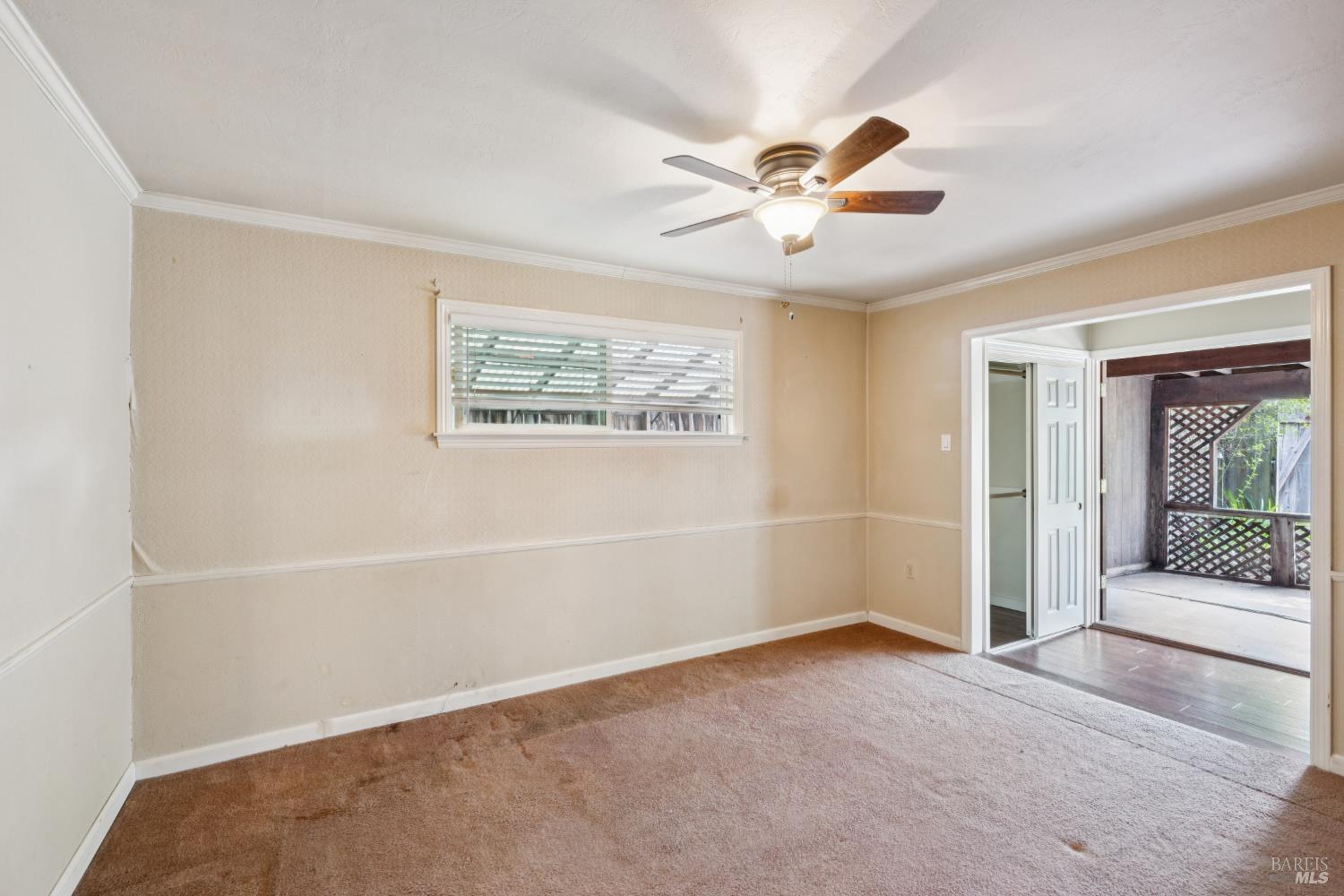 Empty carpeted room with a ceiling fan and window, opening onto a covered patio.